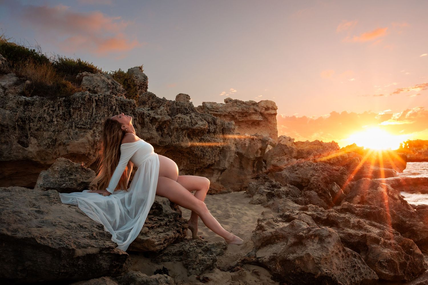 A figure draped in white fabric reclines on coastal rocks at sunset while golden sunlight streams through the cliffs.
