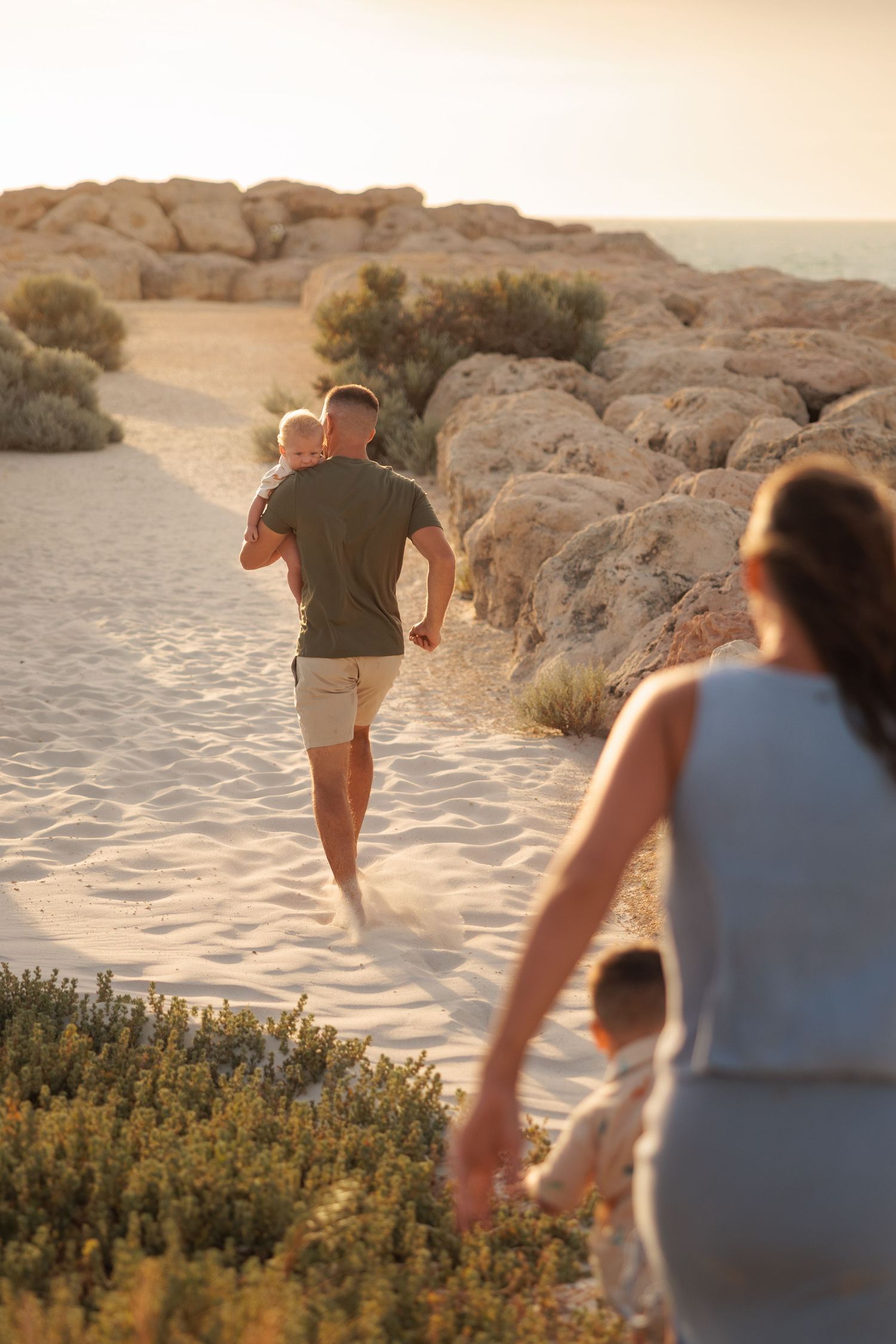 Family walking along sandy beach path at sunset with rocky cliffs and desert vegetation in background.
