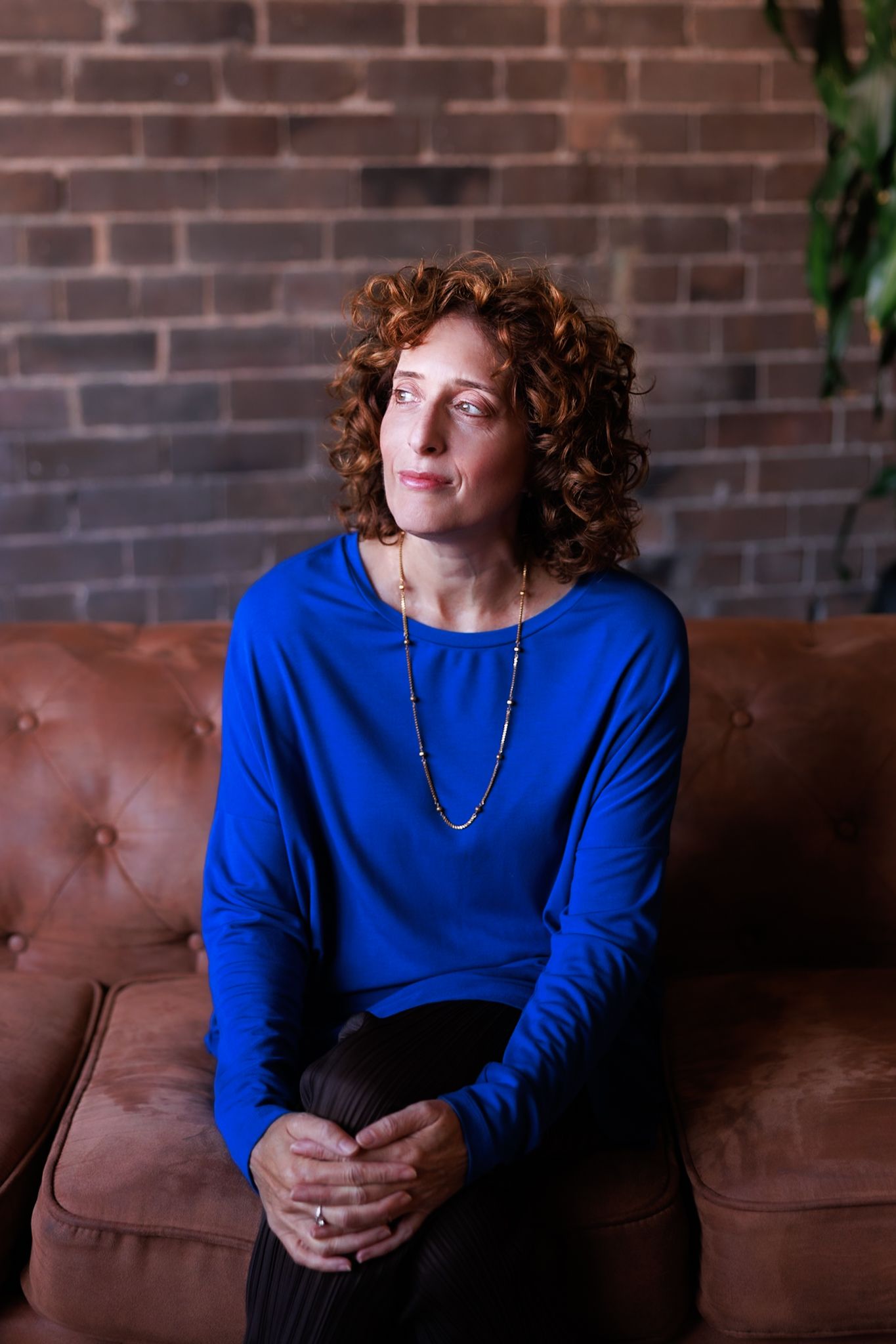 A figure in a vibrant blue sweater poses thoughtfully on a brown leather couch against a brick wall backdrop.