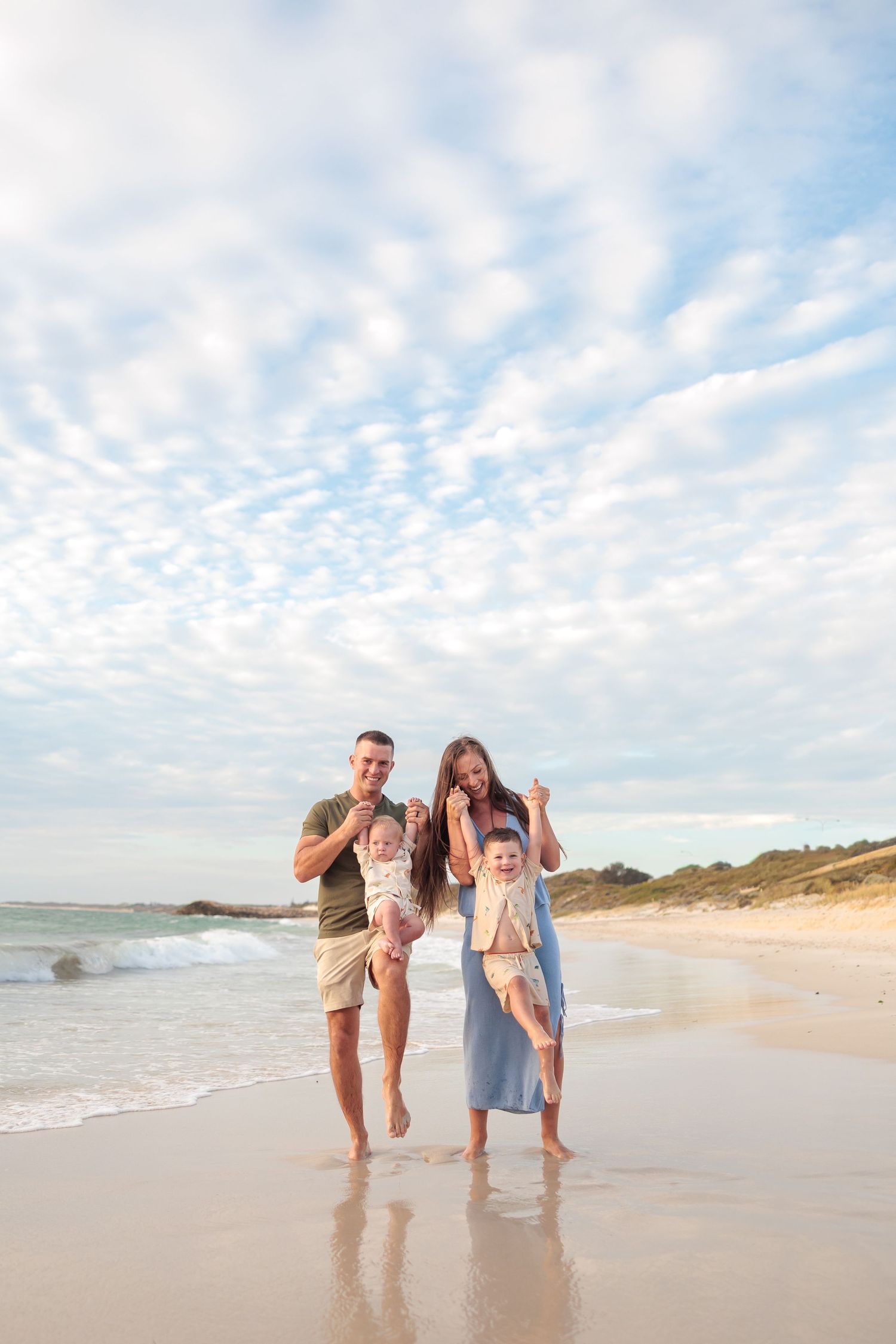 A family in light summer clothing walks and plays together along a sunny beach shoreline with waves and clouds in view.