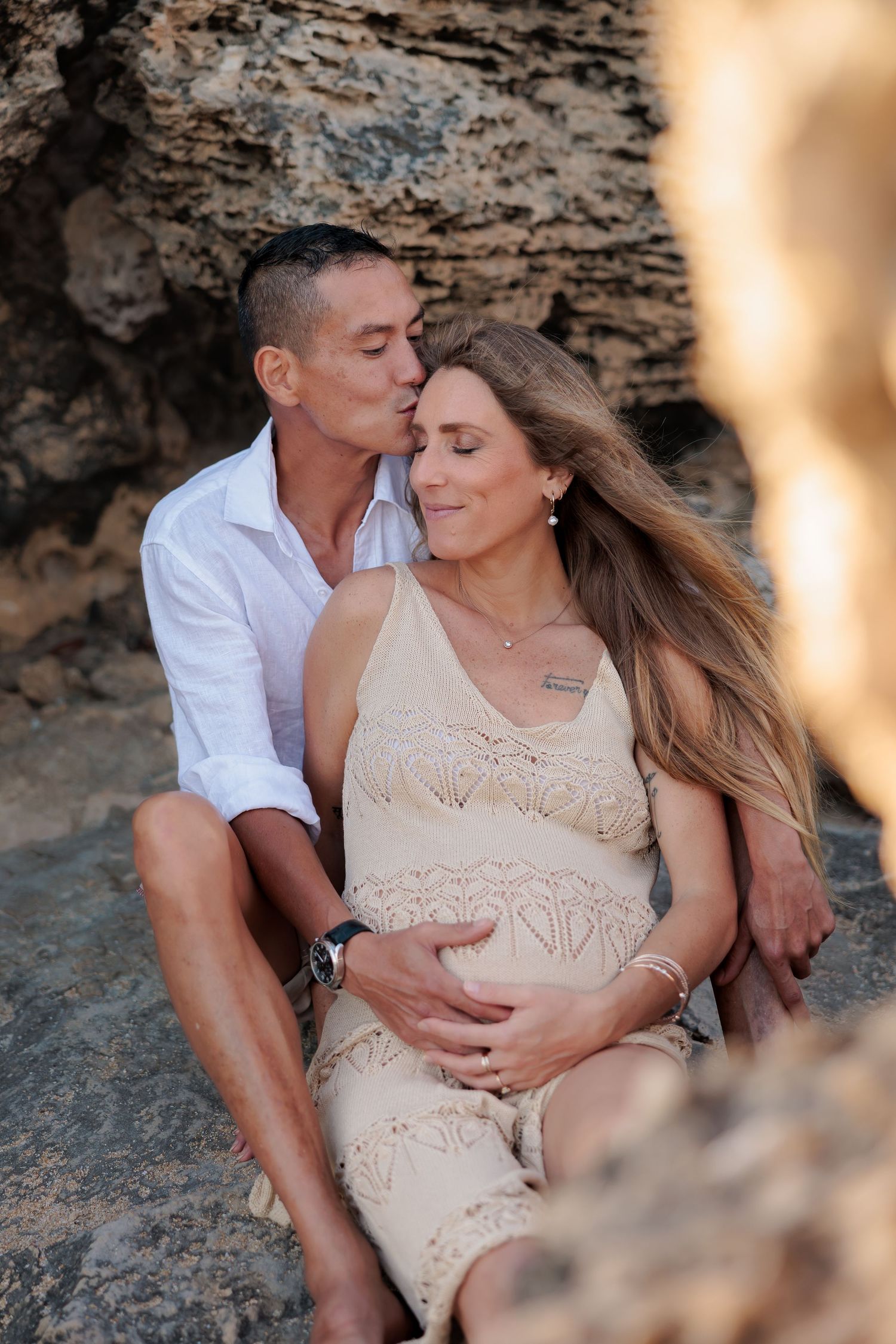 A romantic couple sits together against a rocky cliff wall at sunset in matching beige and white outfits.