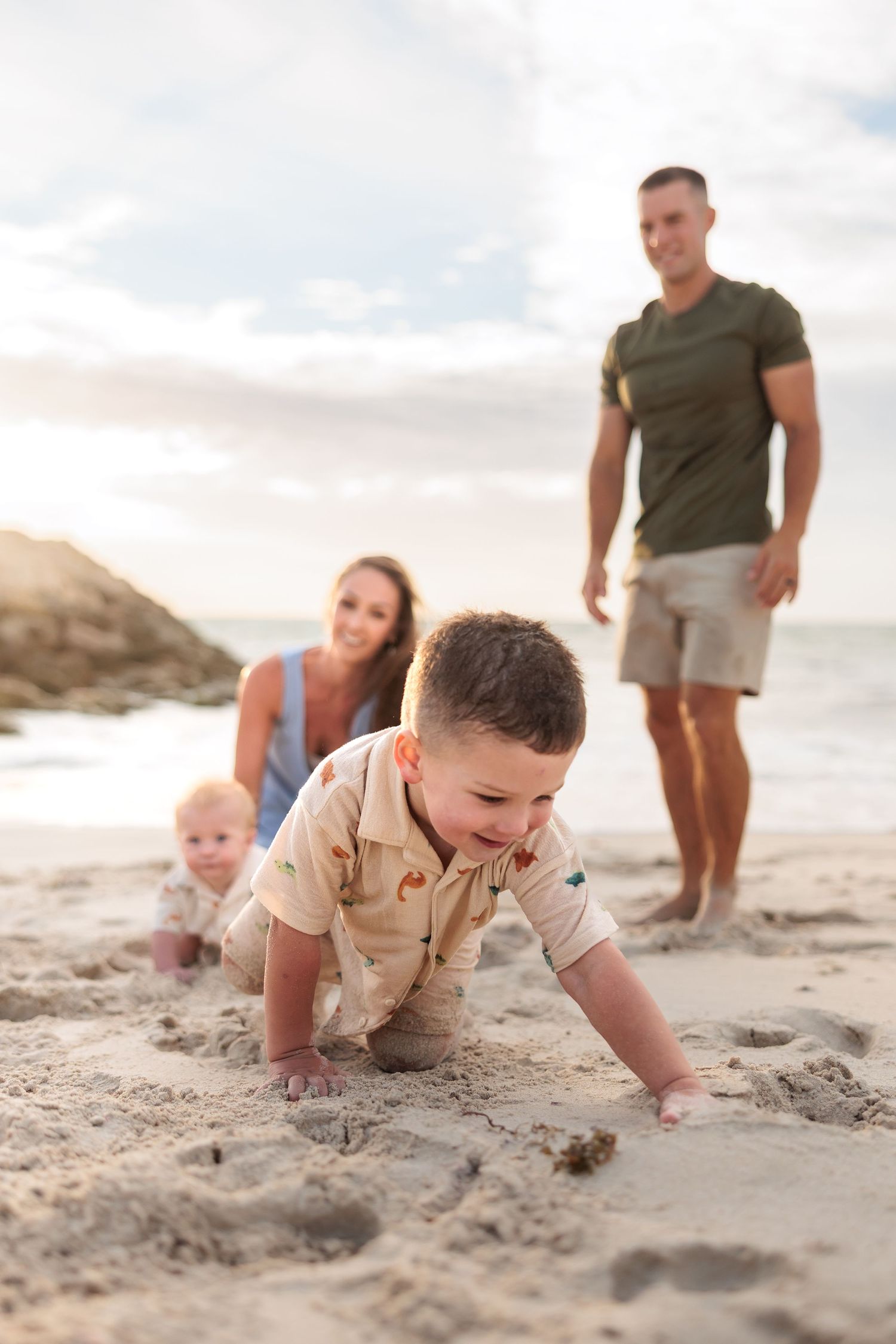 Young child crawls and plays in soft beach sand while others watch from behind on a sunny evening.