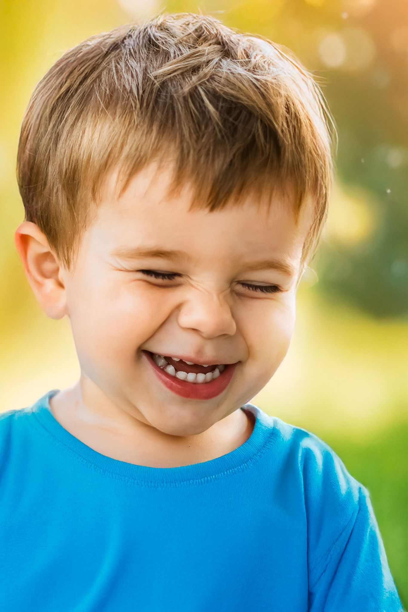 A young child in a blue shirt smiles brightly against a warm sunlit outdoor background.