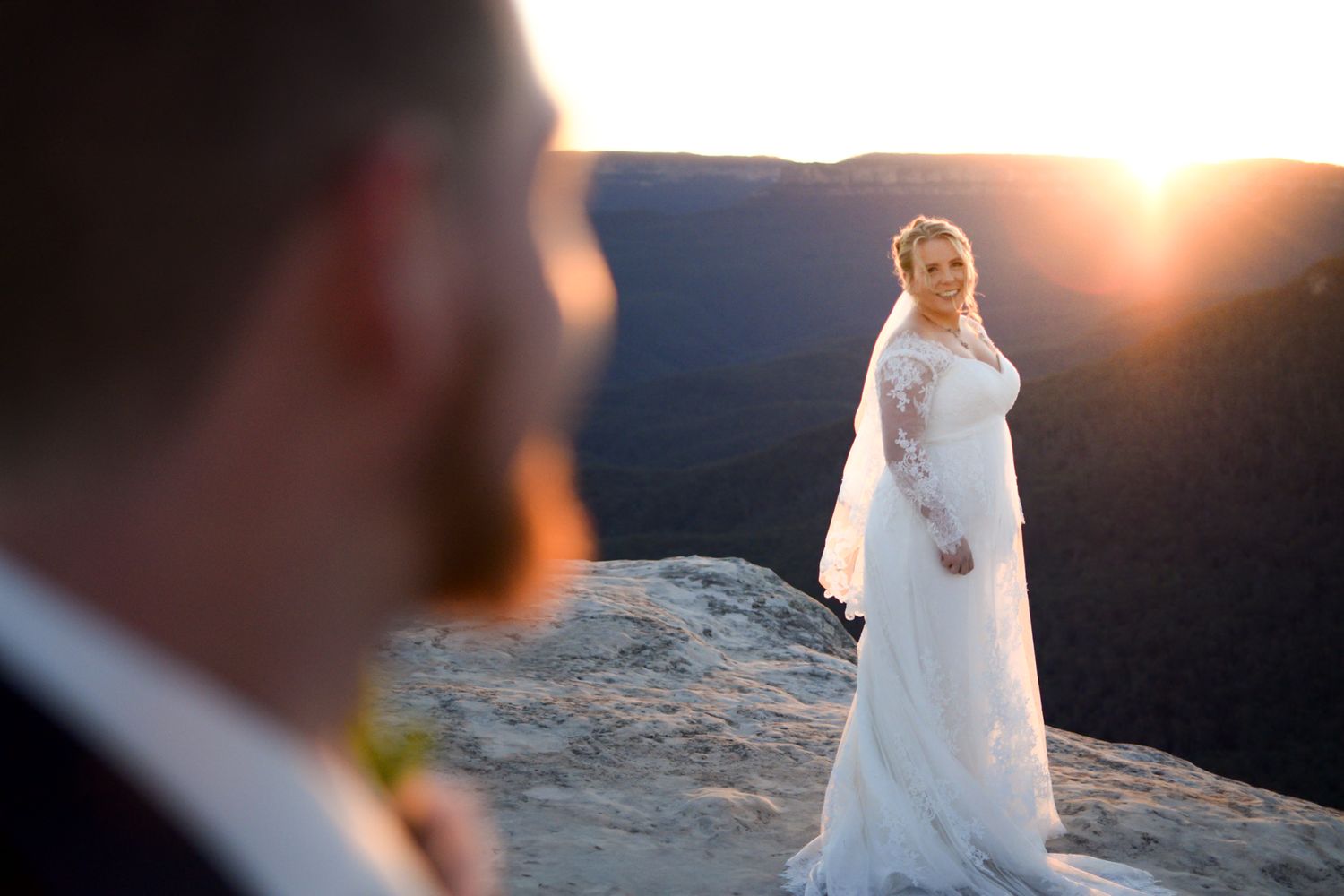 A bride in a white lace wedding dress stands on a cliff edge at sunset while being photographed.