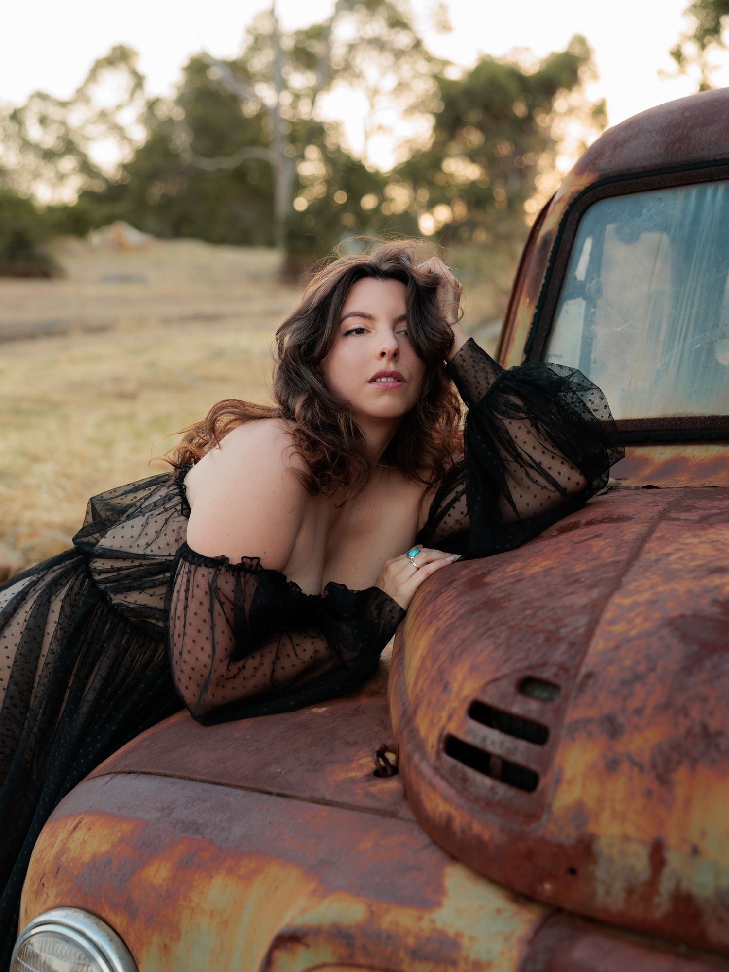 A figure in black lace reclining against a rustic vintage truck in golden sunlight during sunset.
