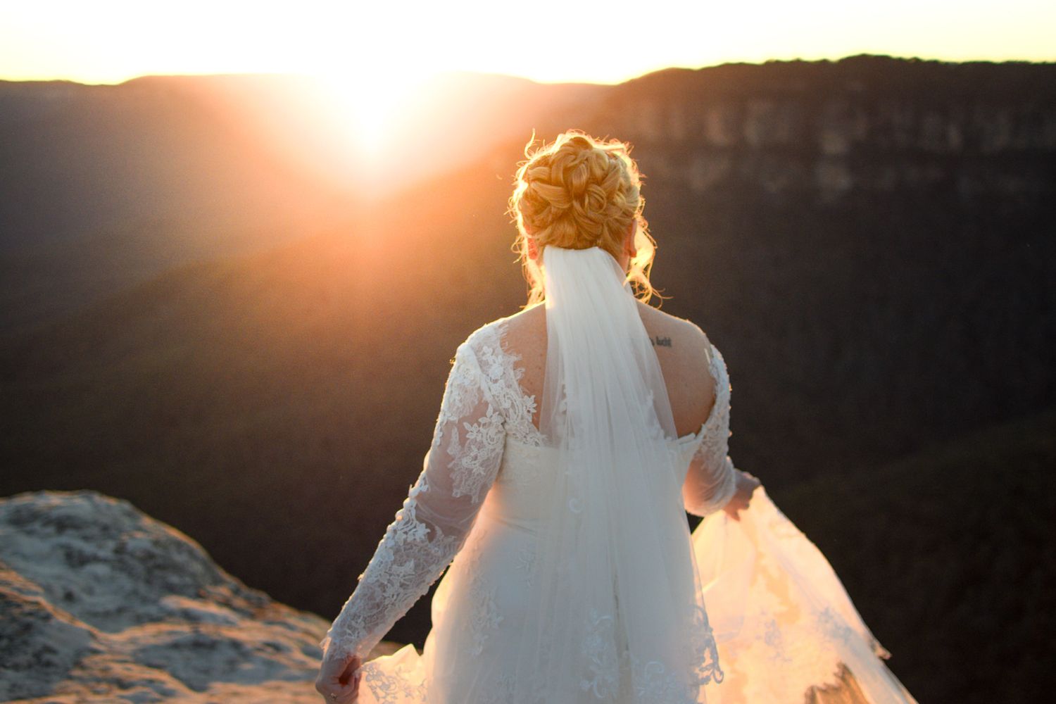 A bride in a lace wedding dress stands at the edge of a canyon during golden hour sunset.