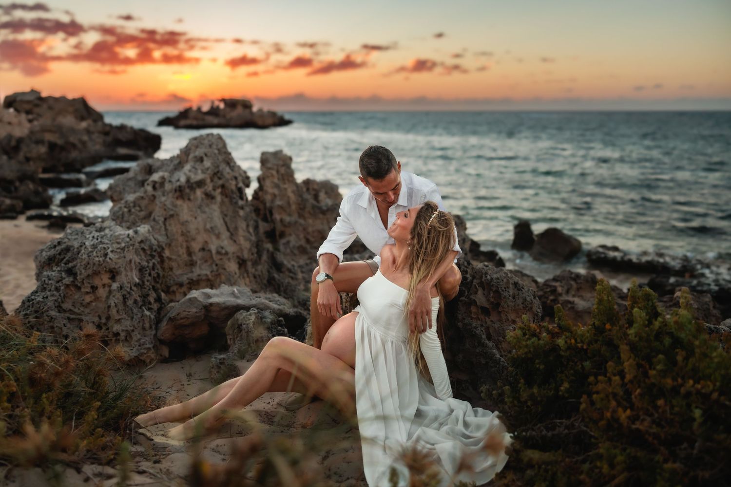 A couple shares a romantic moment on rocky coastal cliffs during a vibrant orange sunset over the ocean.