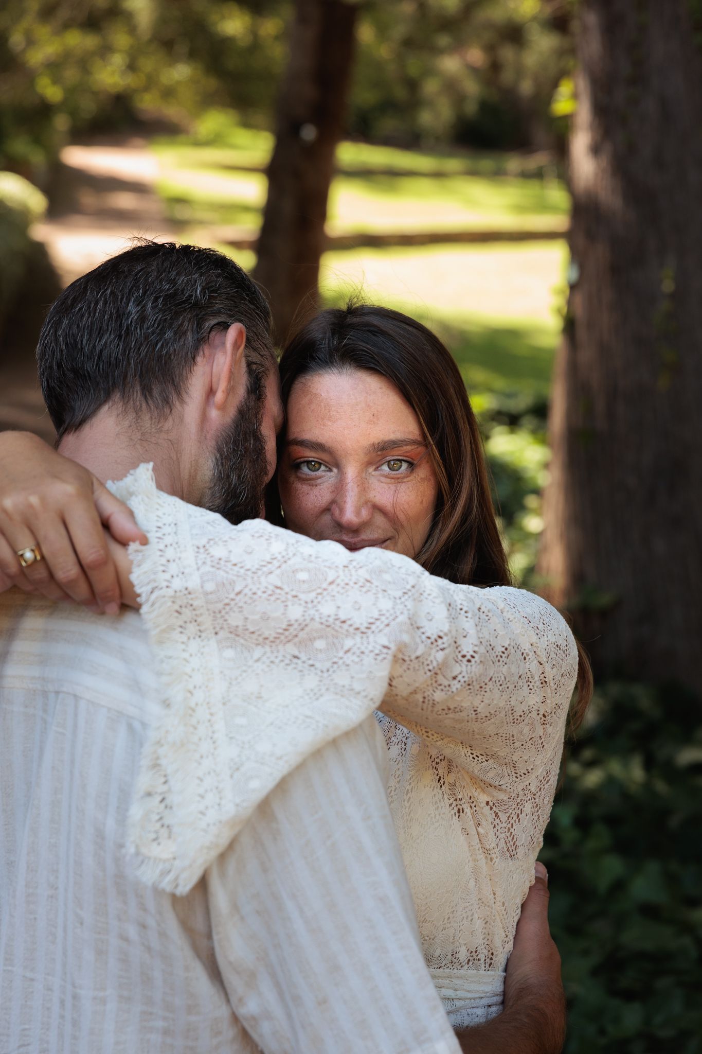 A romantic outdoor embrace between a couple wearing white lace and light colored clothing in a sunlit garden setting.
