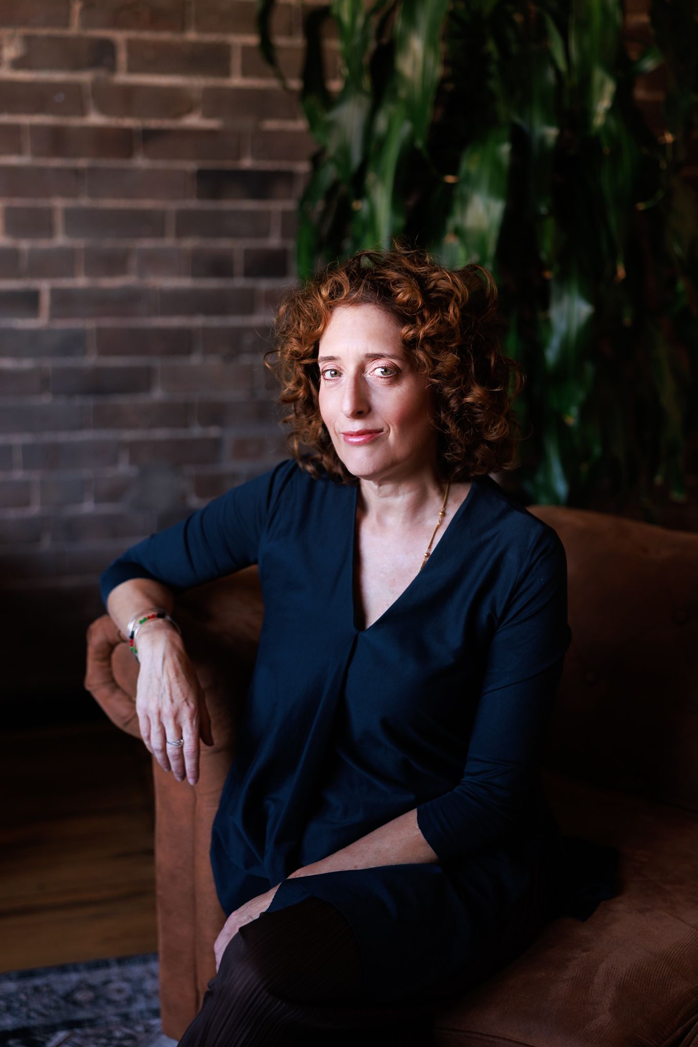 A person with curly red hair wearing a navy blue top poses thoughtfully against a brick wall background.