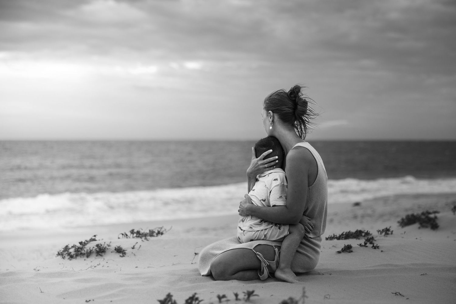 A silhouette sits on a sandy beach looking out at the ocean waves in black and white photography.