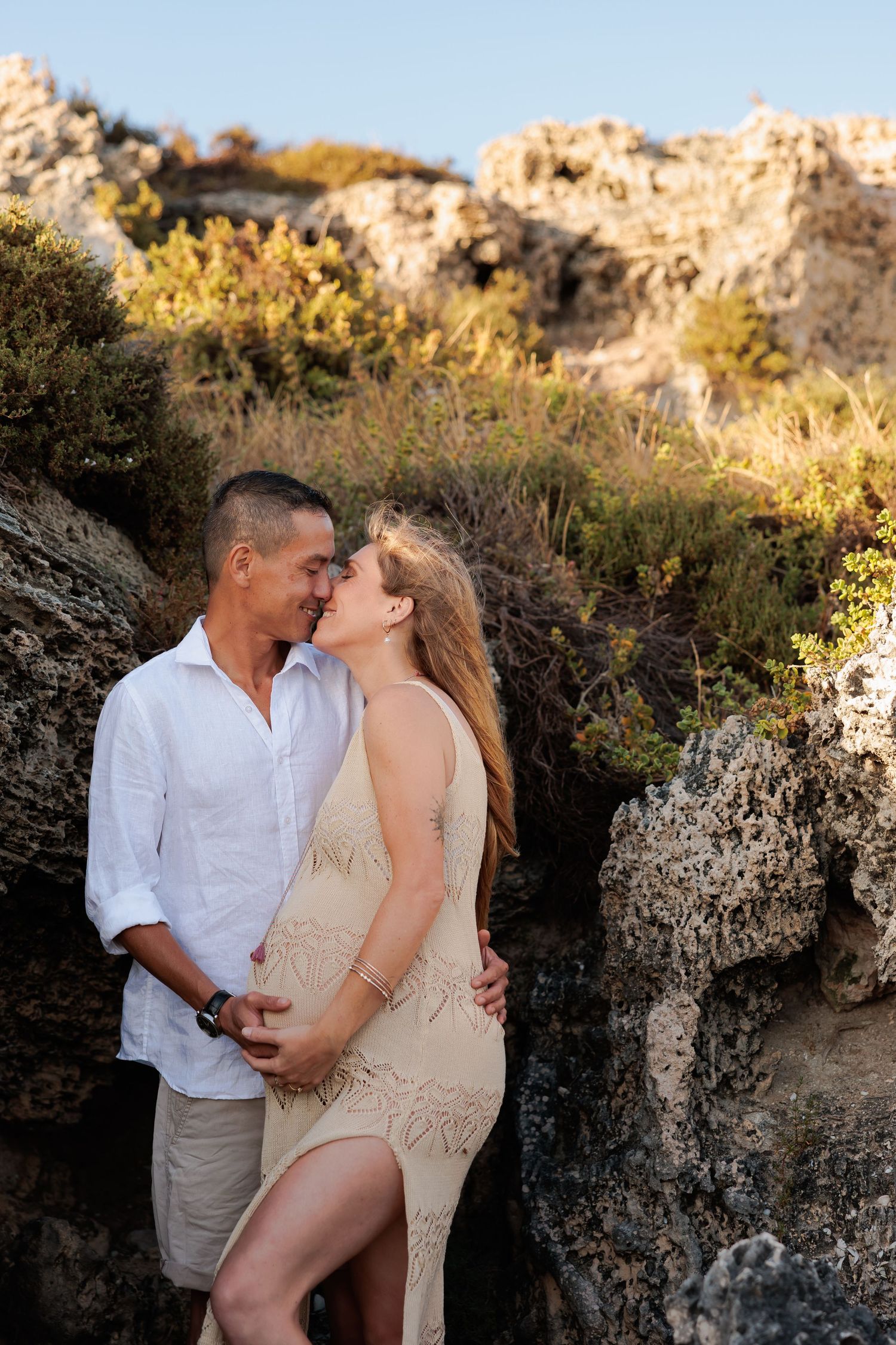 A couple shares an intimate moment against a rocky coastal backdrop during golden hour.