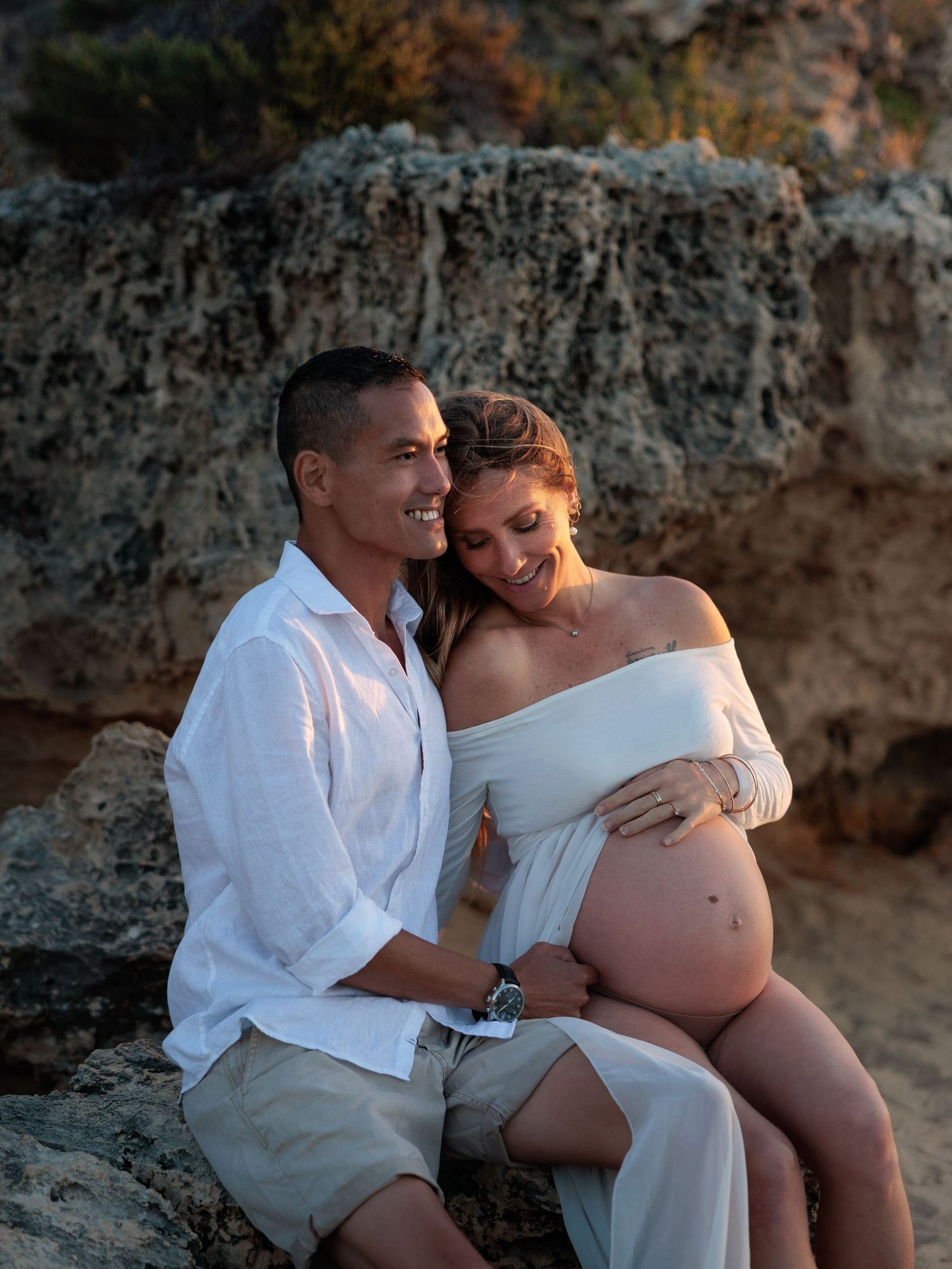 A couple in white clothing shares intimate moments on a beach during a maternity photoshoot at sunset.