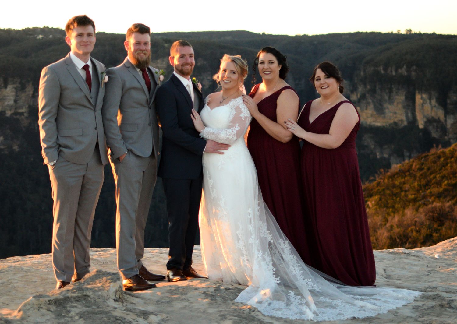 Wedding party stands together against scenic mountain backdrop at sunset in formal burgundy and gray attire.