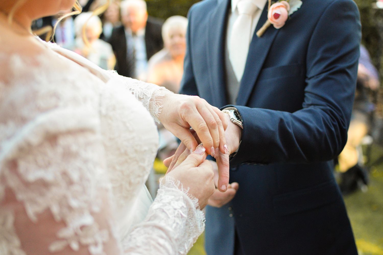 Close-up of hands exchanging wedding rings during an outdoor ceremony in natural sunlight.