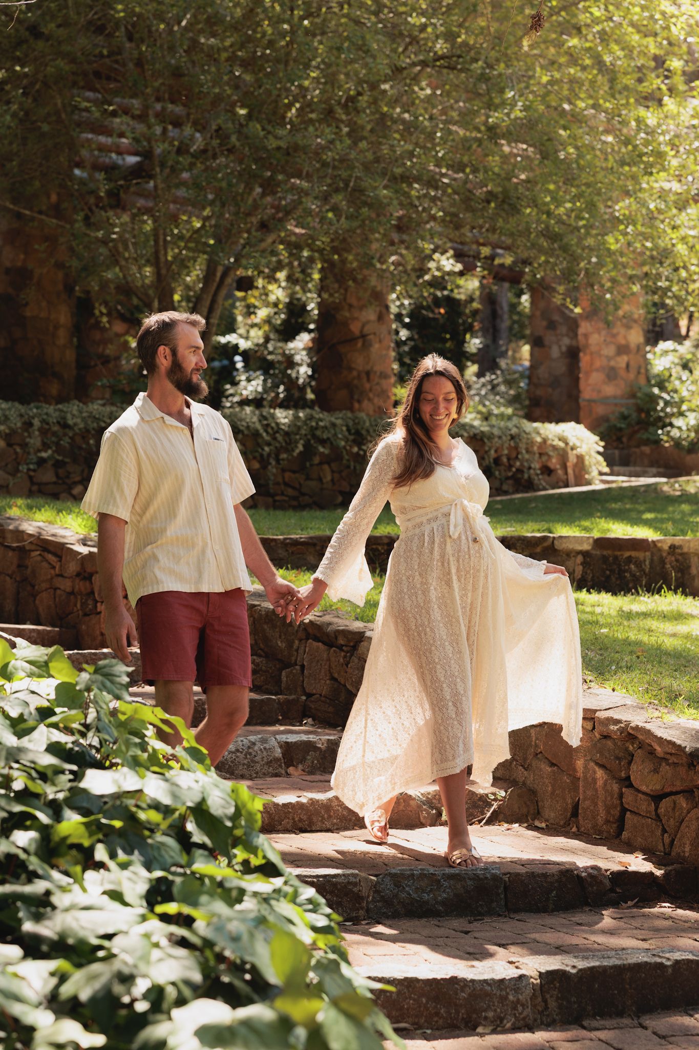 A couple in white outfits sits together on rustic stone steps surrounded by lush greenery in a garden setting.