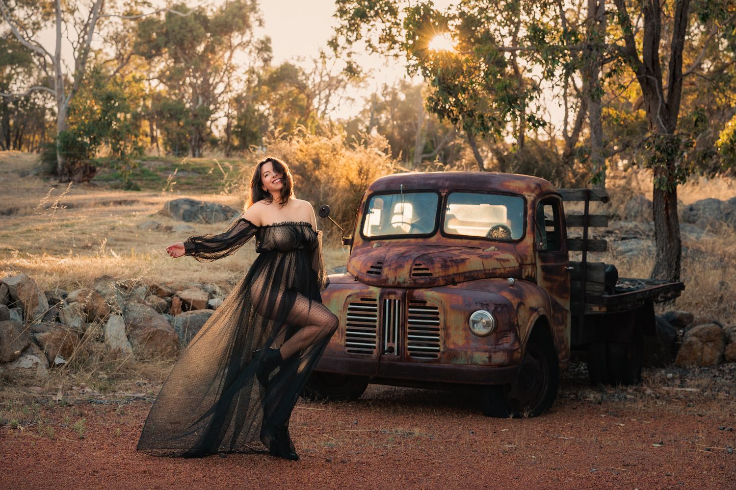 A vintage rusty car sits in a golden field at sunset while someone in a flowing dress poses near it.