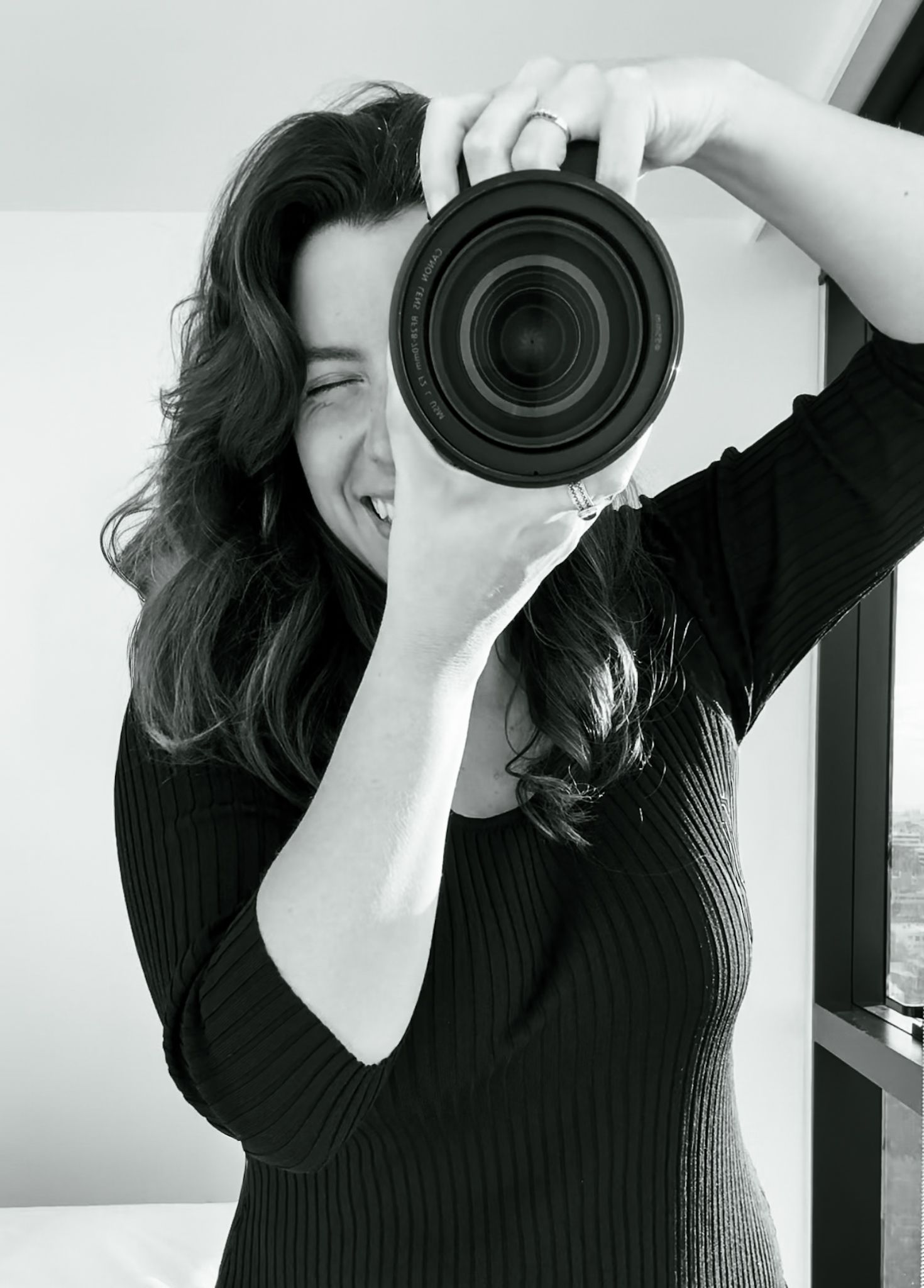 A joyful photographer in a black sweater holds up a professional camera lens while taking a picture in black and white.