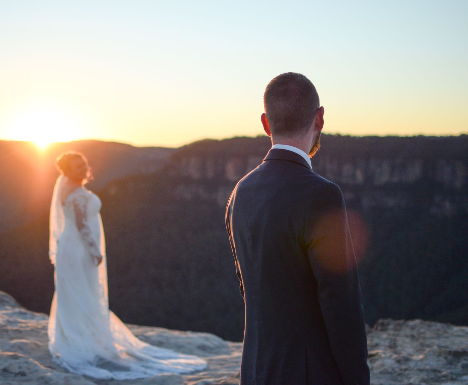 Newlywed couple stands at scenic mountain overlook during golden sunset while bride's veil flows in the breeze.