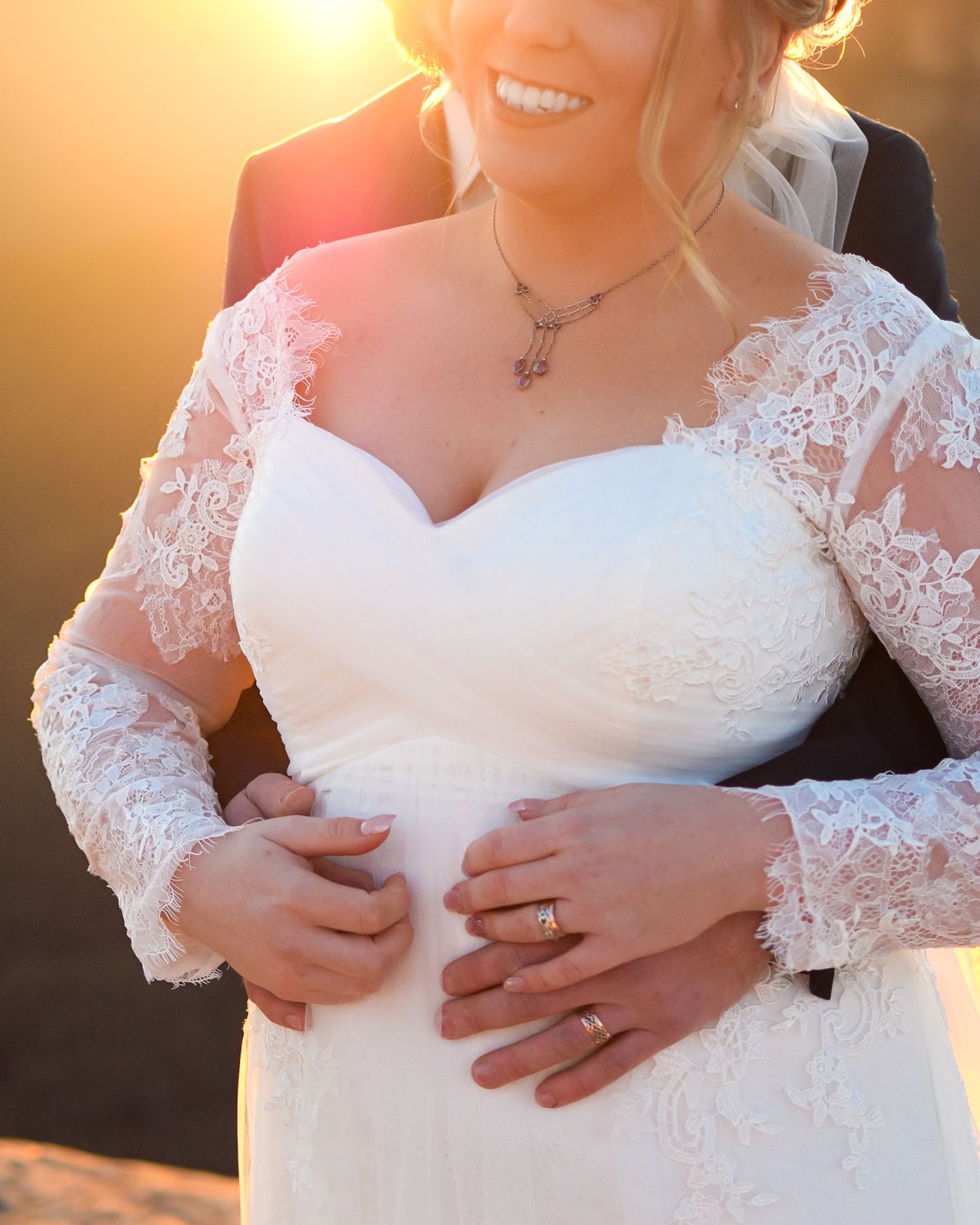 Close-up details of a white lace wedding dress with delicate hand placement showing rings.