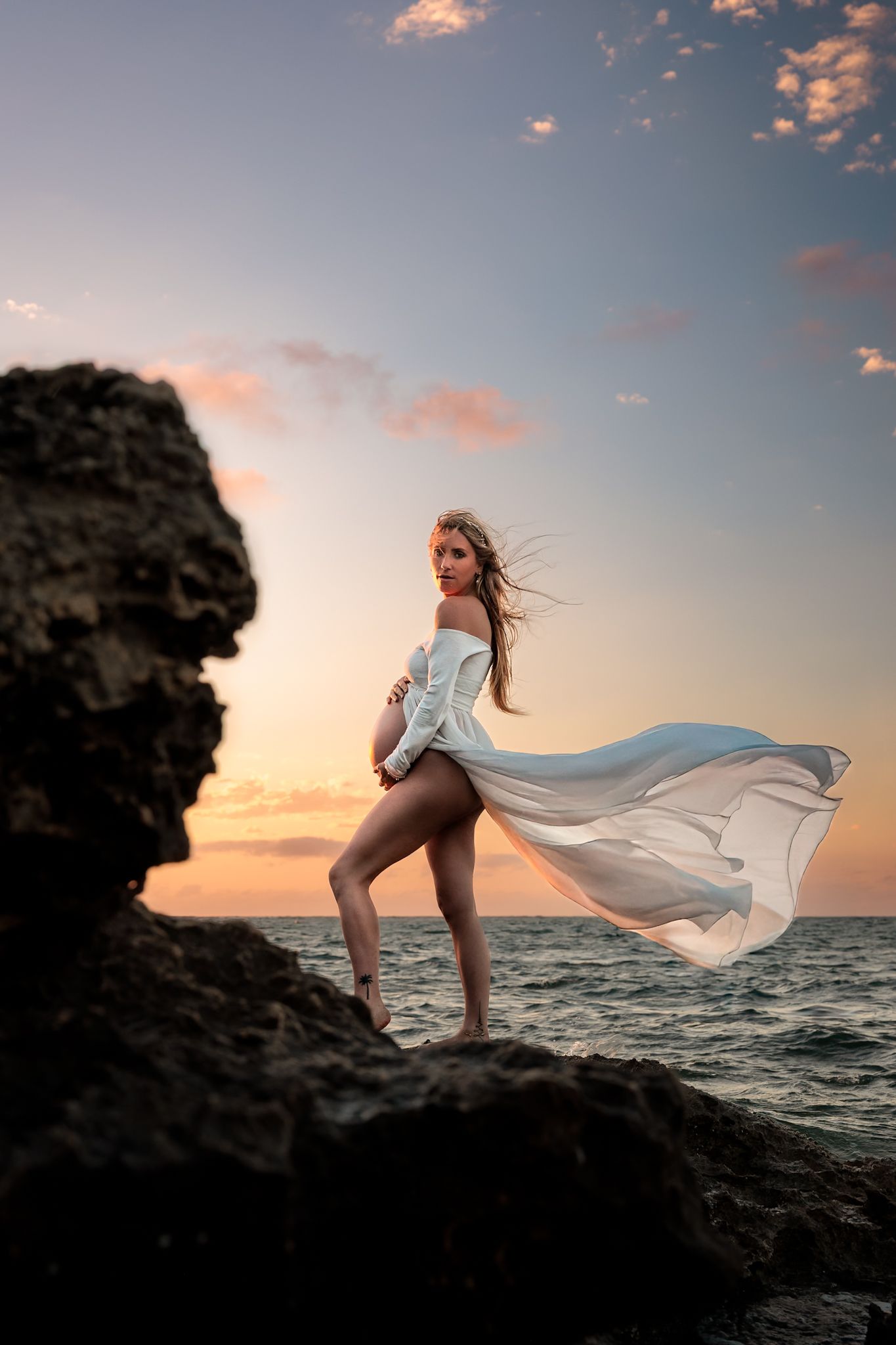A flowing white dress billows dramatically against a rocky coastal sunset backdrop creating an ethereal silhouette.