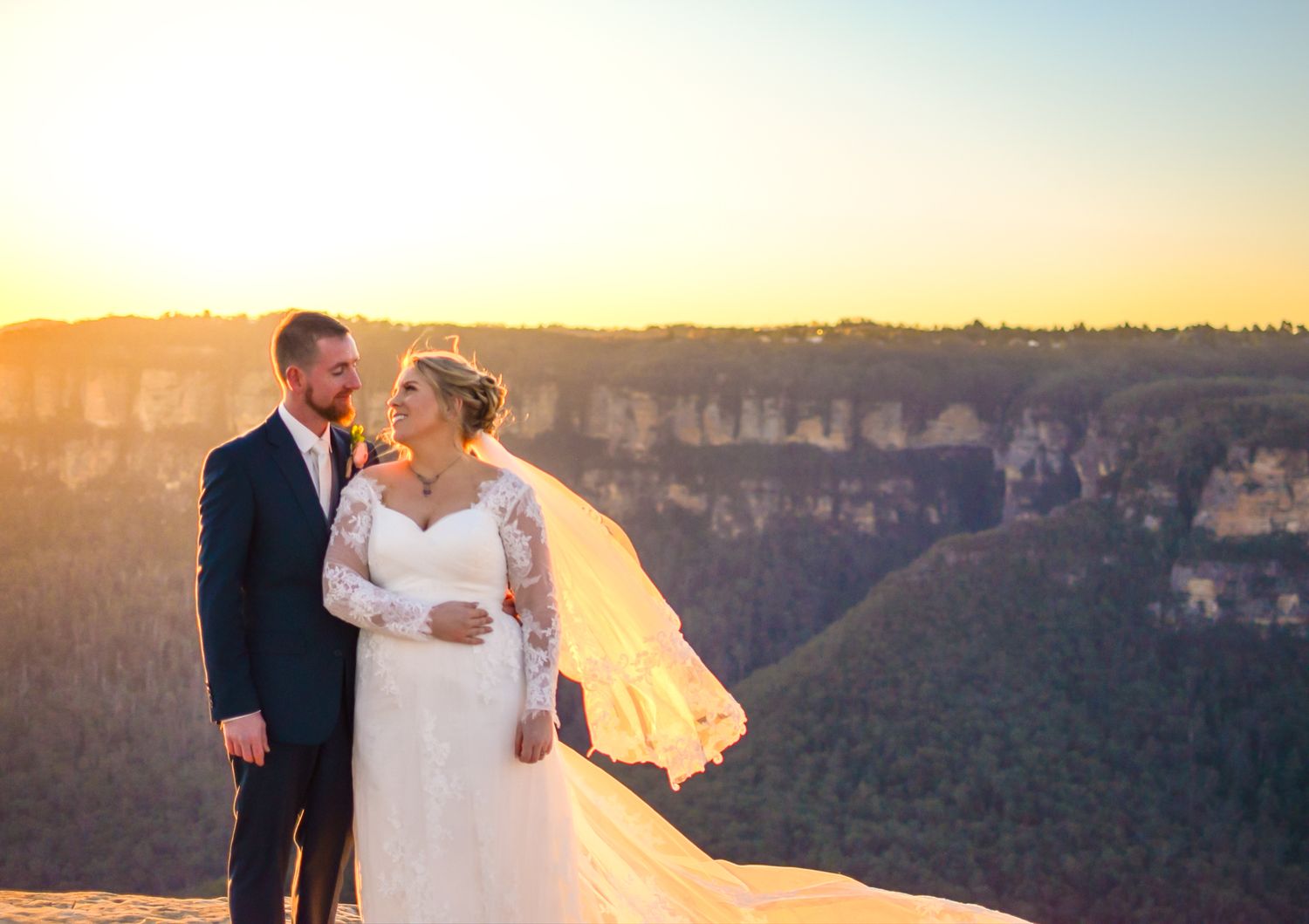 A couple stands silhouetted against a golden sunset at the Grand Canyon, with dramatic cliffs in the background.