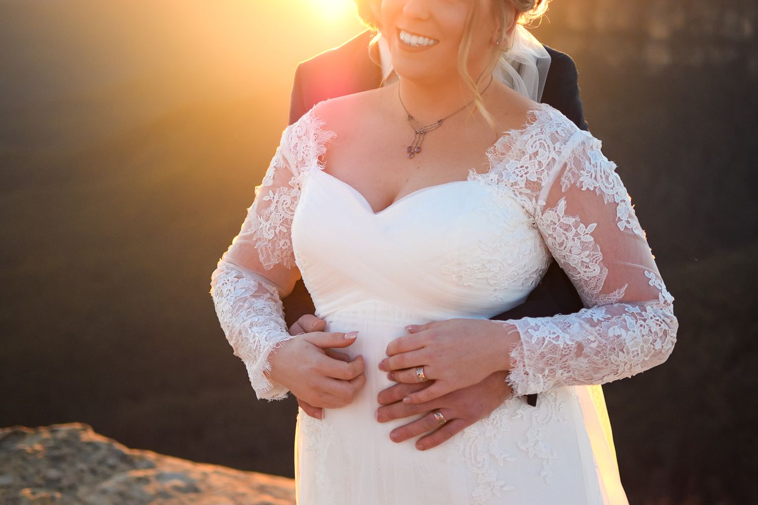 A bride in a white lace wedding dress poses during golden hour with sunlight creating a warm glow in the background.