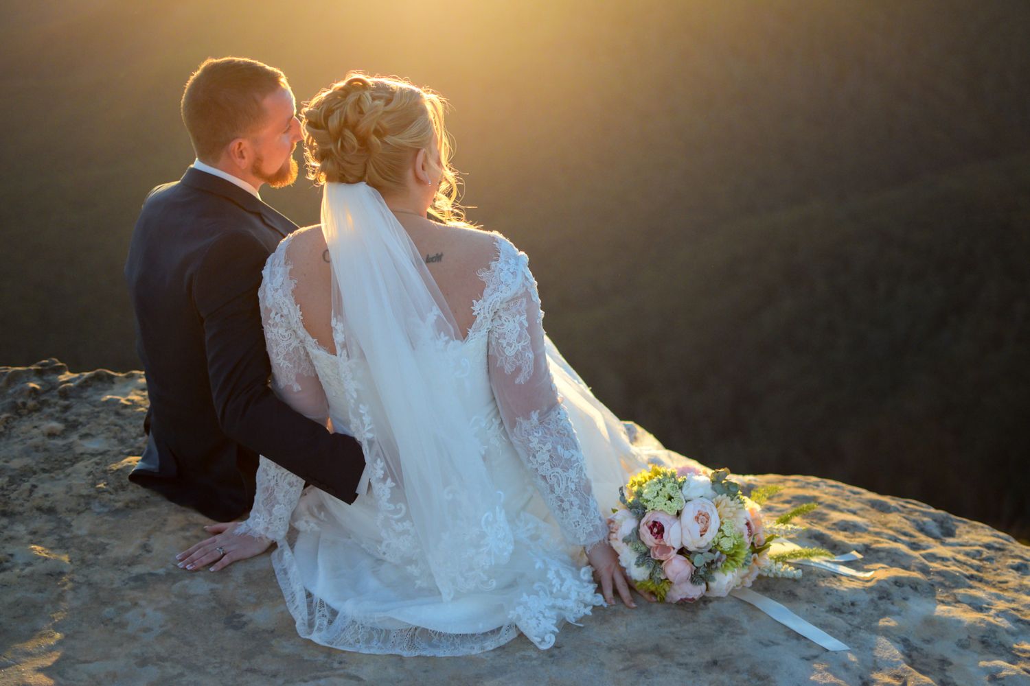 A newlywed couple sits on a cliff edge at sunset with a bridal bouquet beside them in golden light.