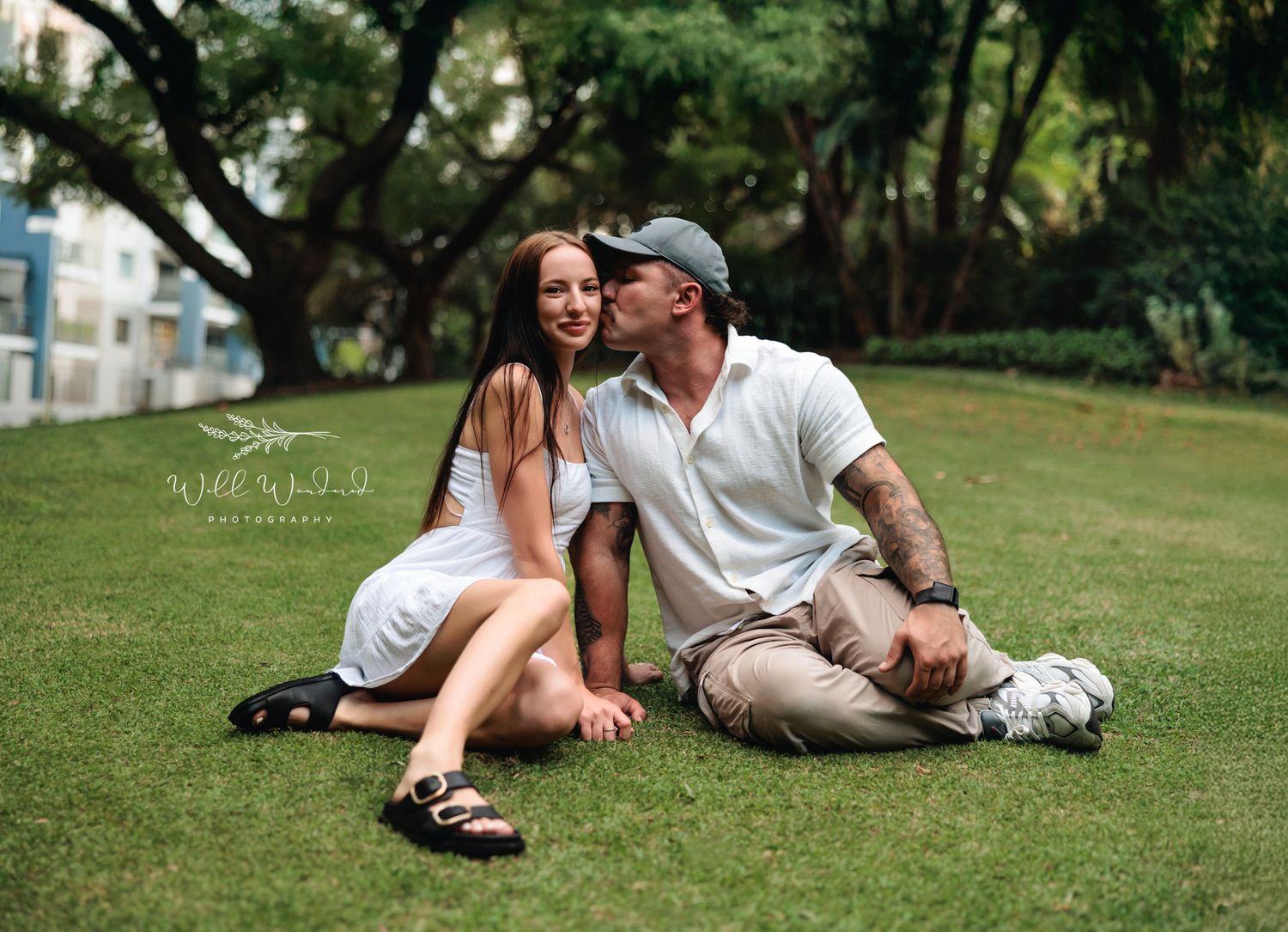 A couple in white and beige casual attire sits together on green grass in a park setting on a sunny day.