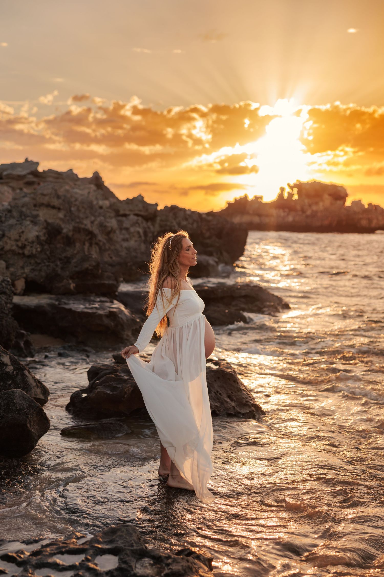 A flowing white dress catches the golden light as waves crash against rocky shores during a dramatic coastal sunset.