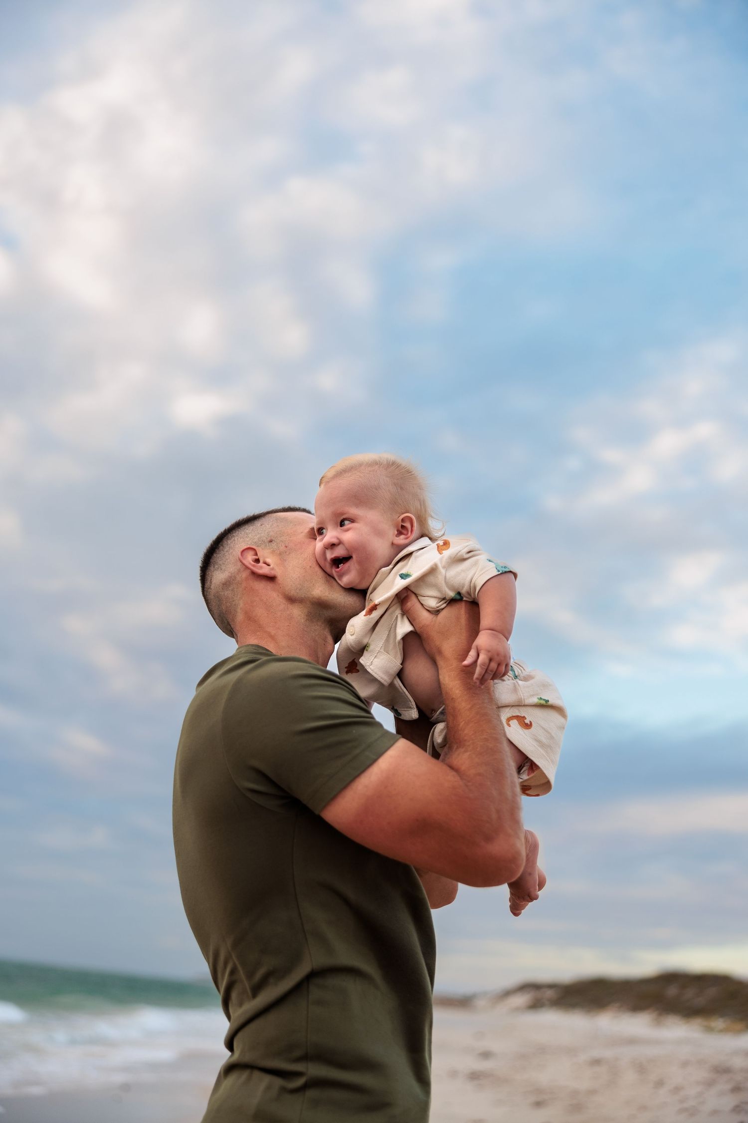 A tender moment shared between an adult and baby on a sunny beach with ocean waves and clouds in the background.