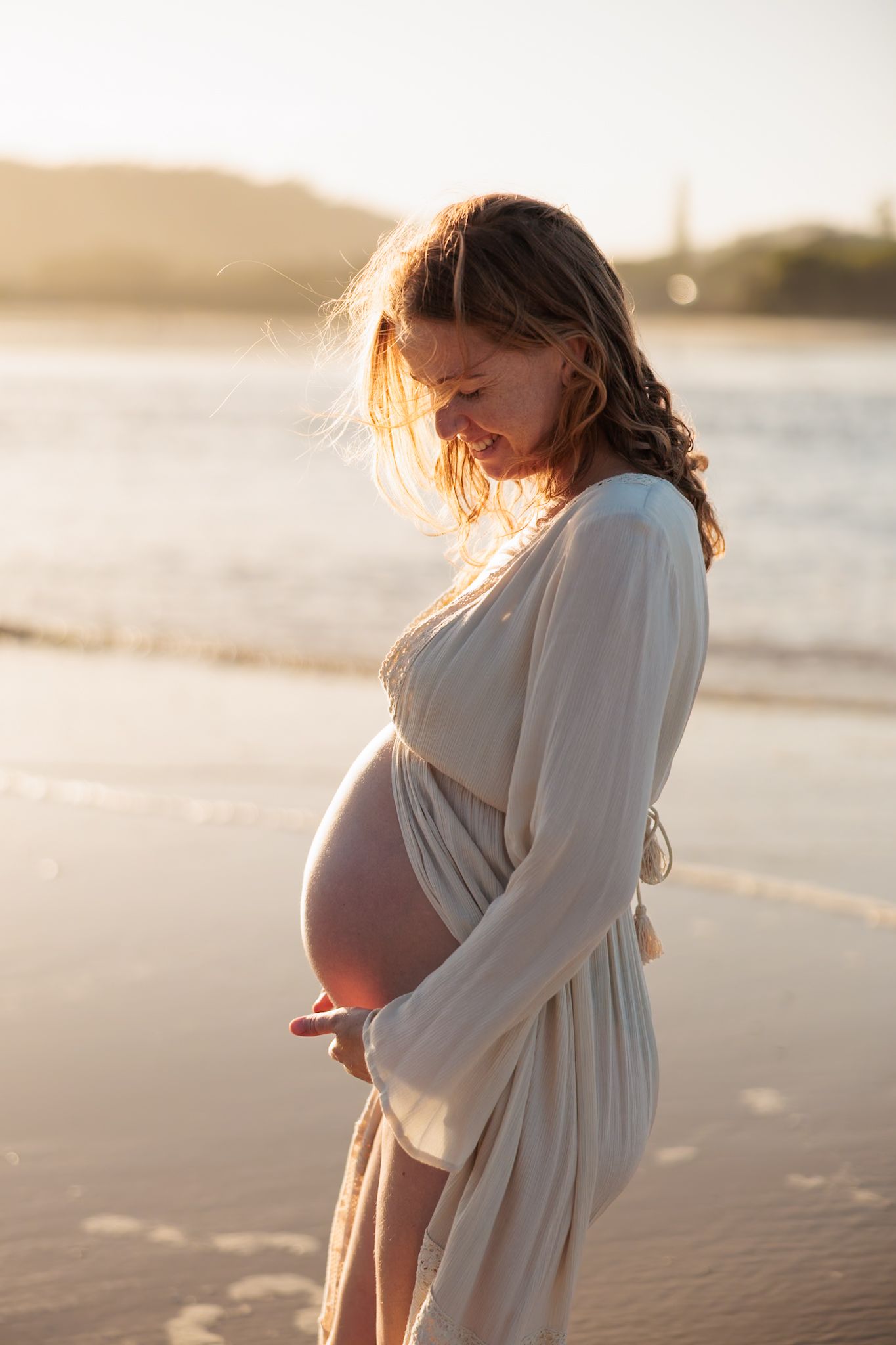A pregnant woman in a white dress stands on a beach during golden hour sunset, silhouetted against the ocean waves.