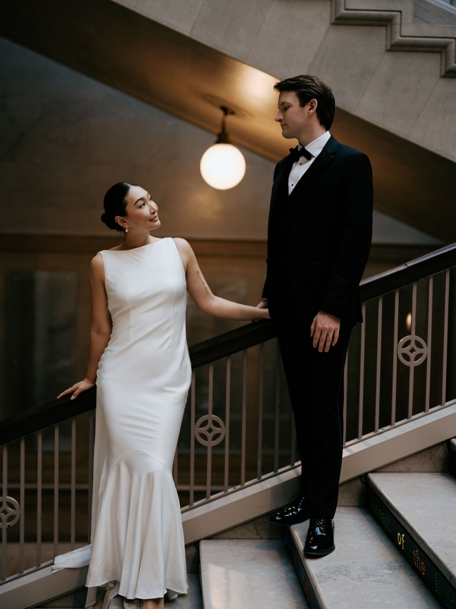 Bride and groom sharing a tender moment on ornate staircase, bride in sleek white gown, groom in tuxedo.