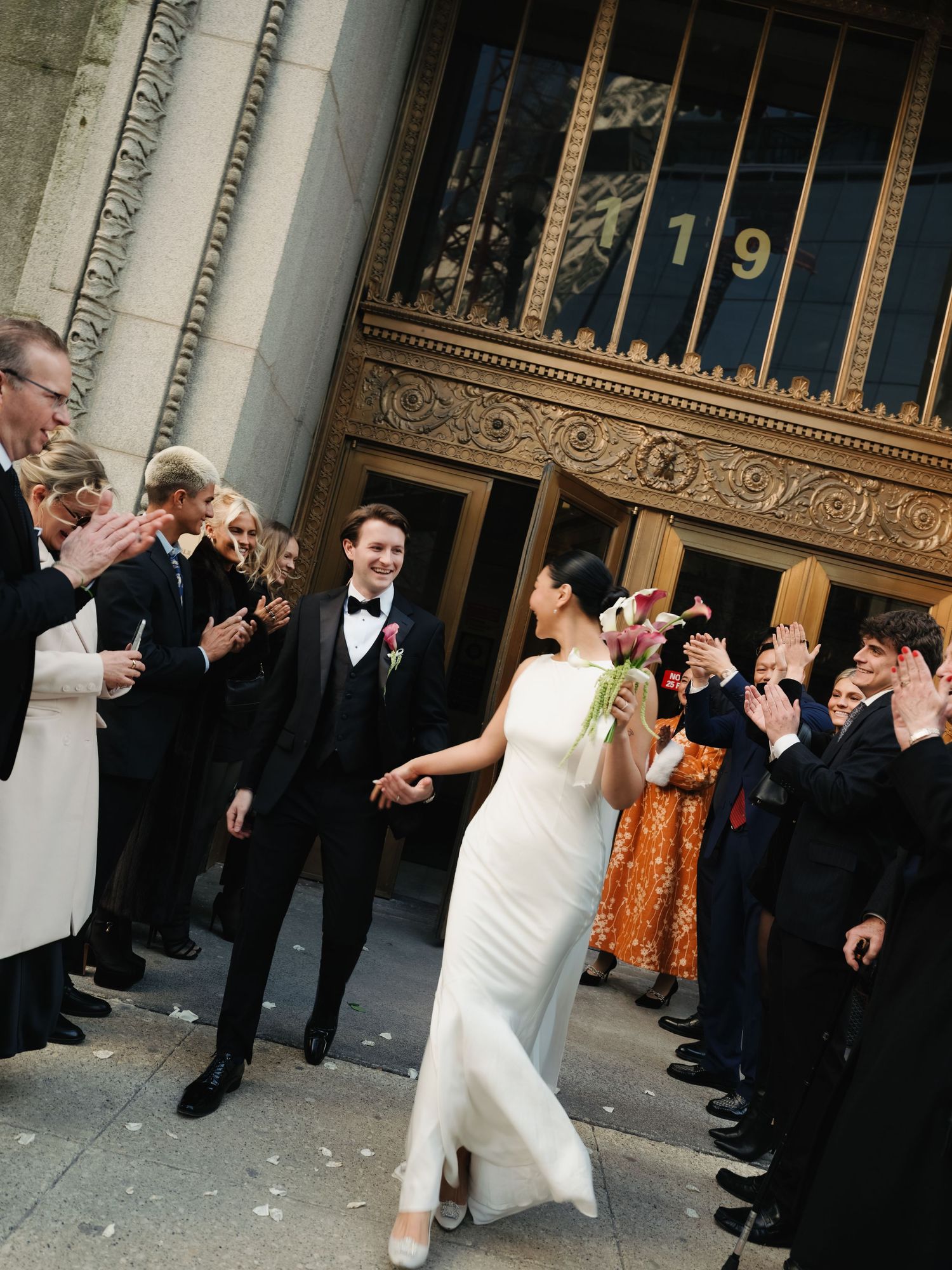 Bride in elegant white gown and groom in black tuxedo hold hands exiting ornate building with guests.