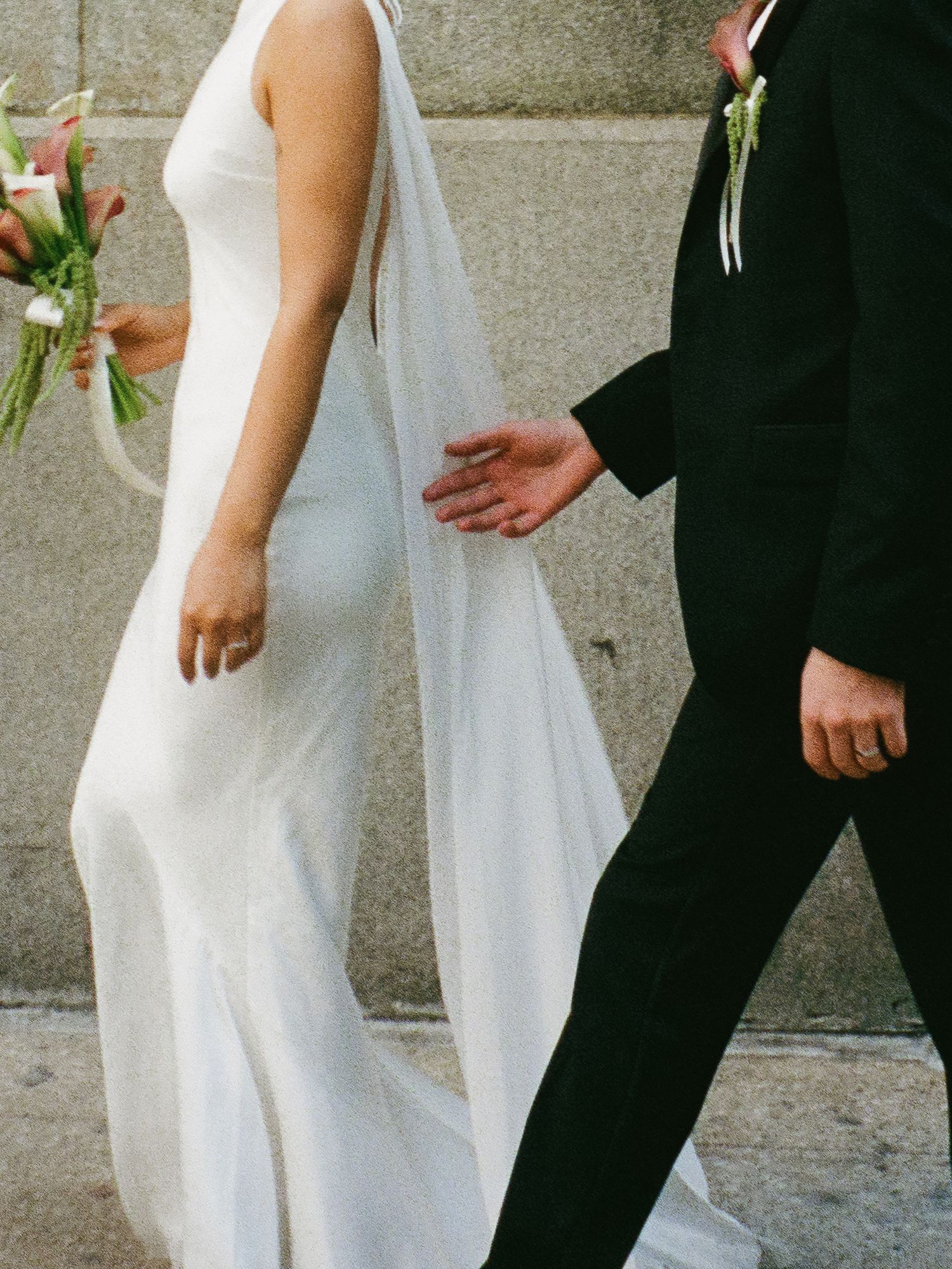 Bride in sleek white gown holds bouquet while walking hand-in-hand with groom in black suit.