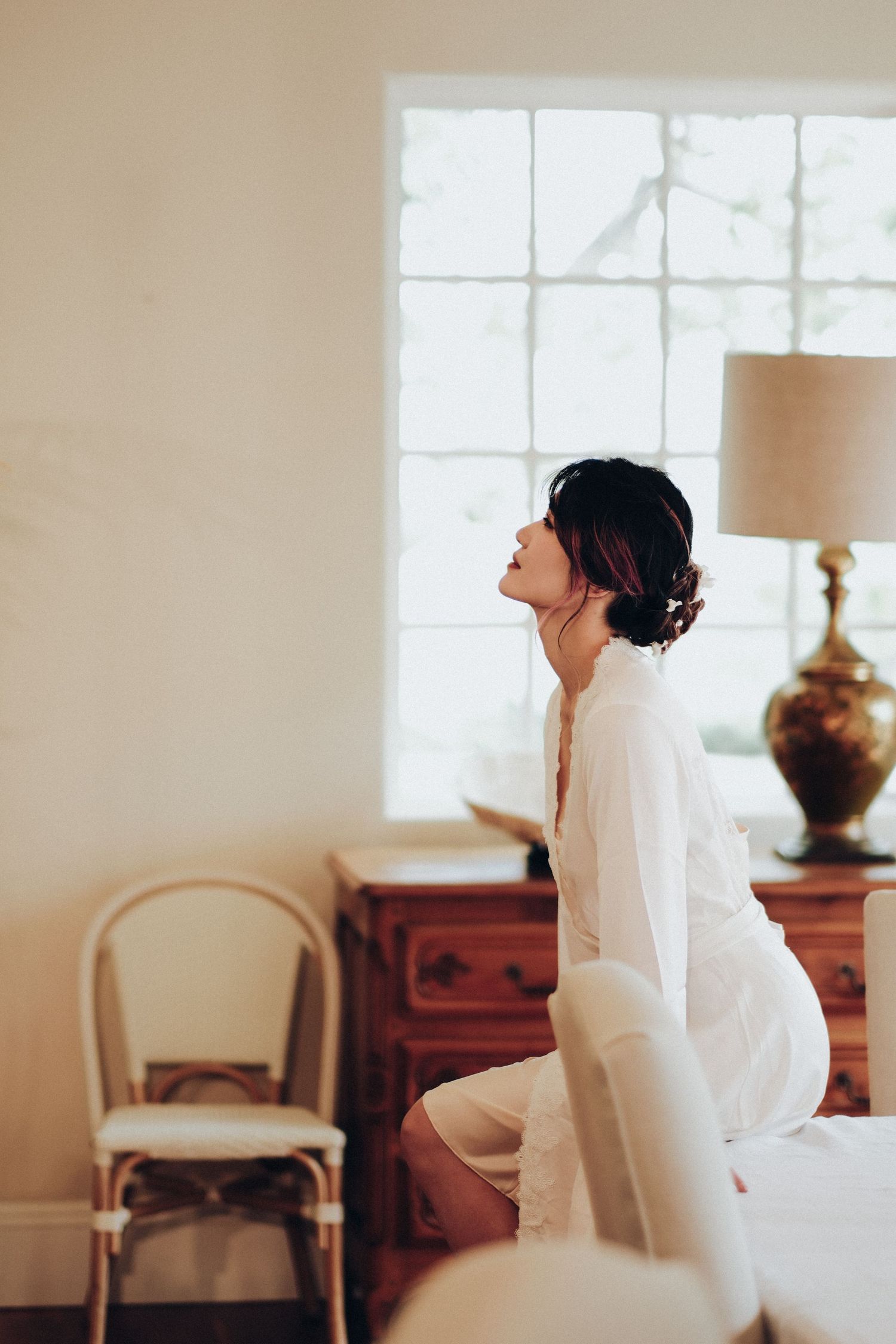 Bride in white robe sitting on bed gazing upward in softly lit wedding morning scene.