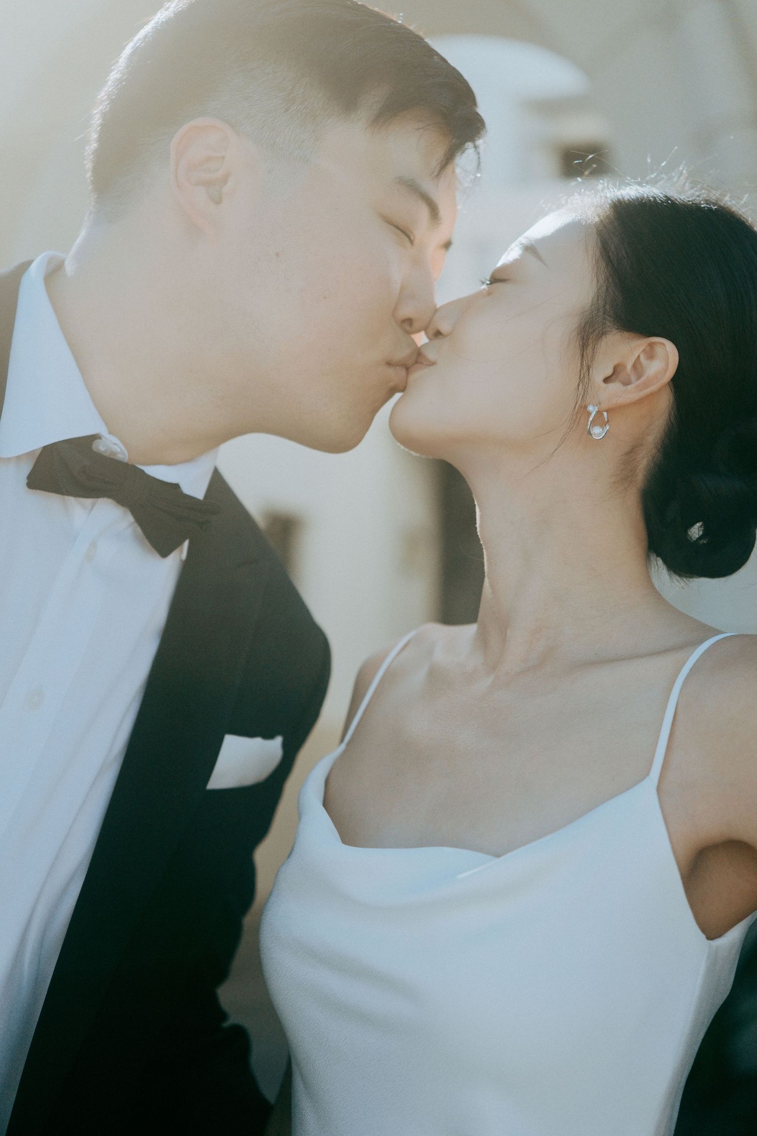 Asian bride and groom sharing a romantic kiss outdoors in soft sunlight on their wedding day.