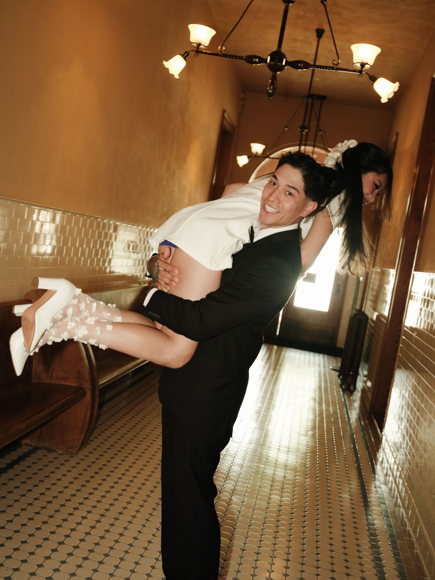 Groom joyfully carries bride in white mini dress through sunlit tiled hallway, both laughing.