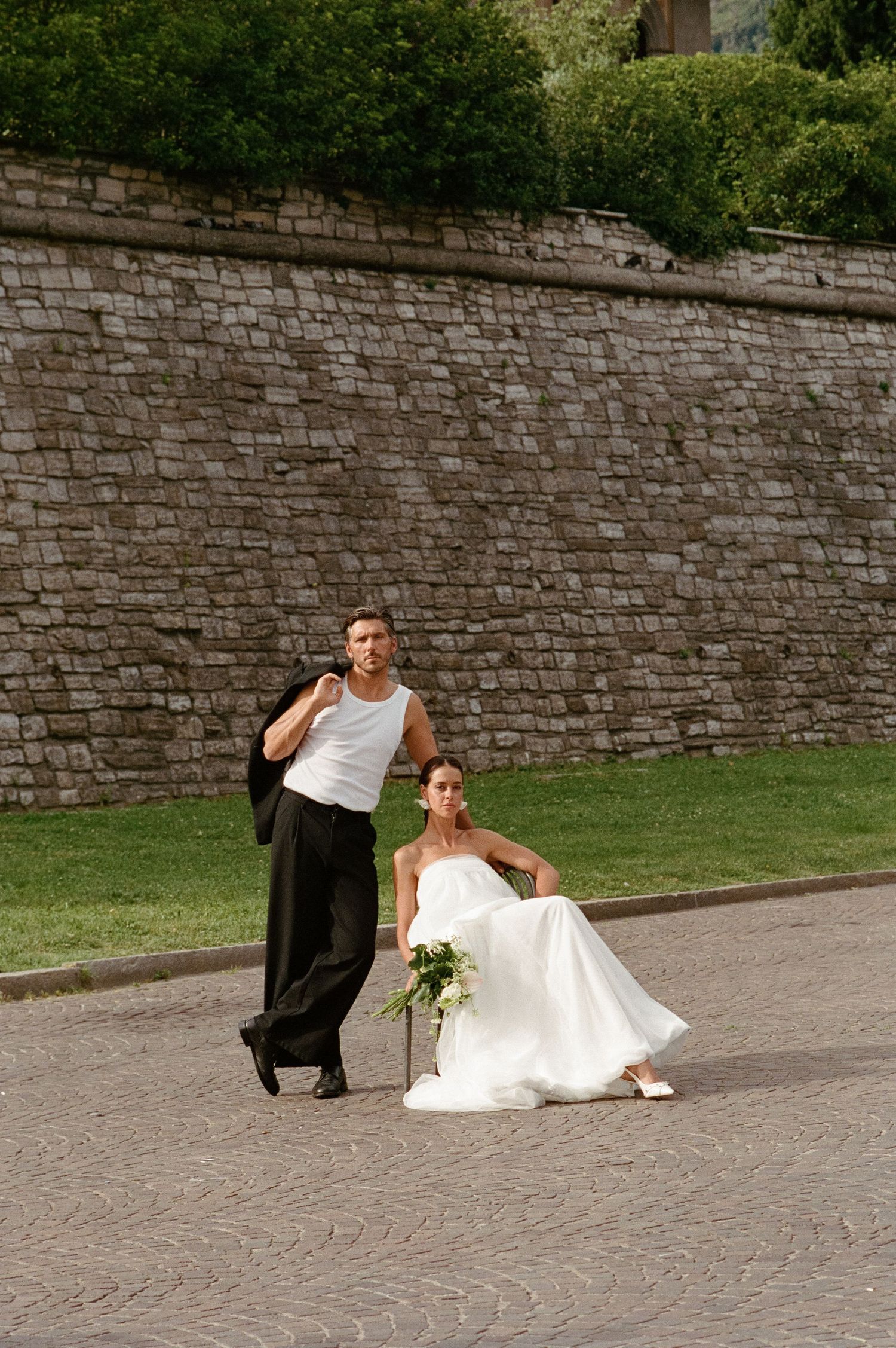 Bride seated on cobblestones in white gown holding green bouquet beside groom in white tank top.