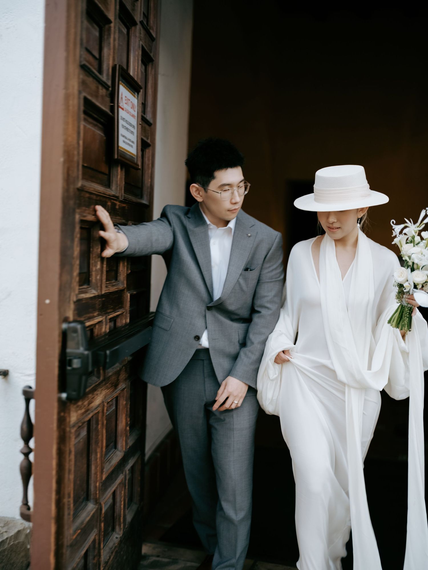Bride in white gown and hat exits historic wooden door with groom in gray suit.
