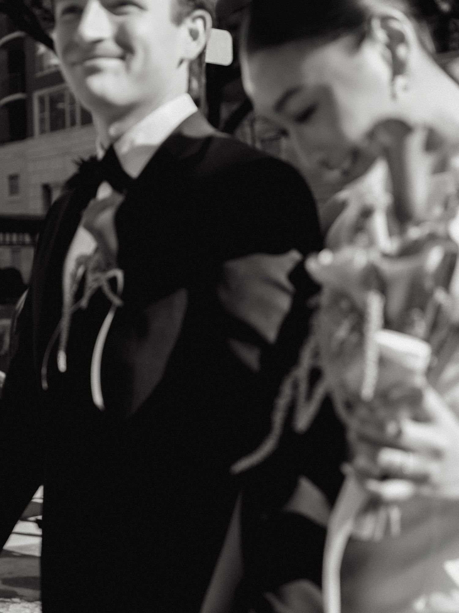 Black and white photo of a bride and groom walking outdoors, groom in tuxedo, bride holding flowers.
