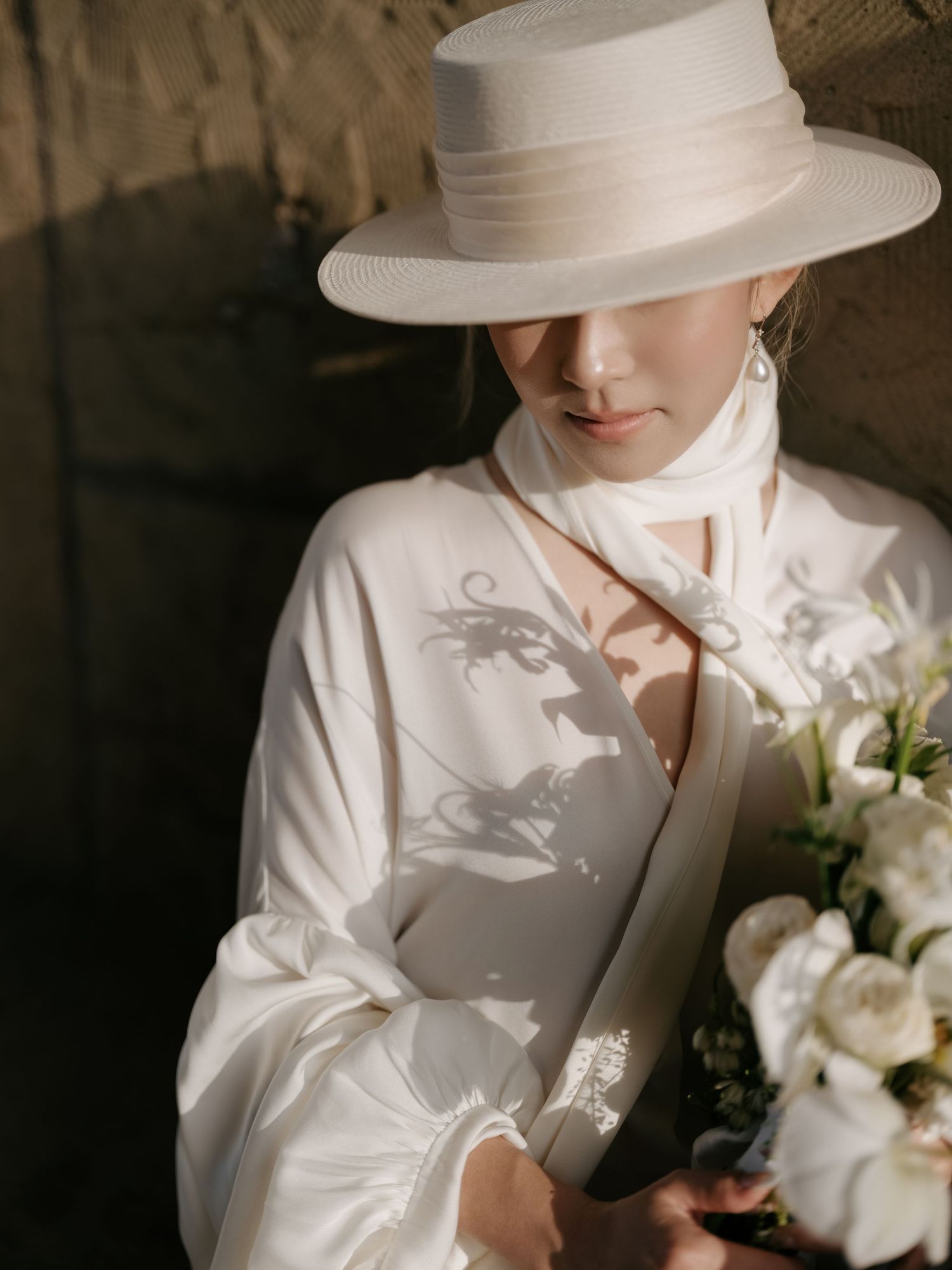 Bride in white wide-brim hat and flowing dress holds white floral bouquet in soft light.