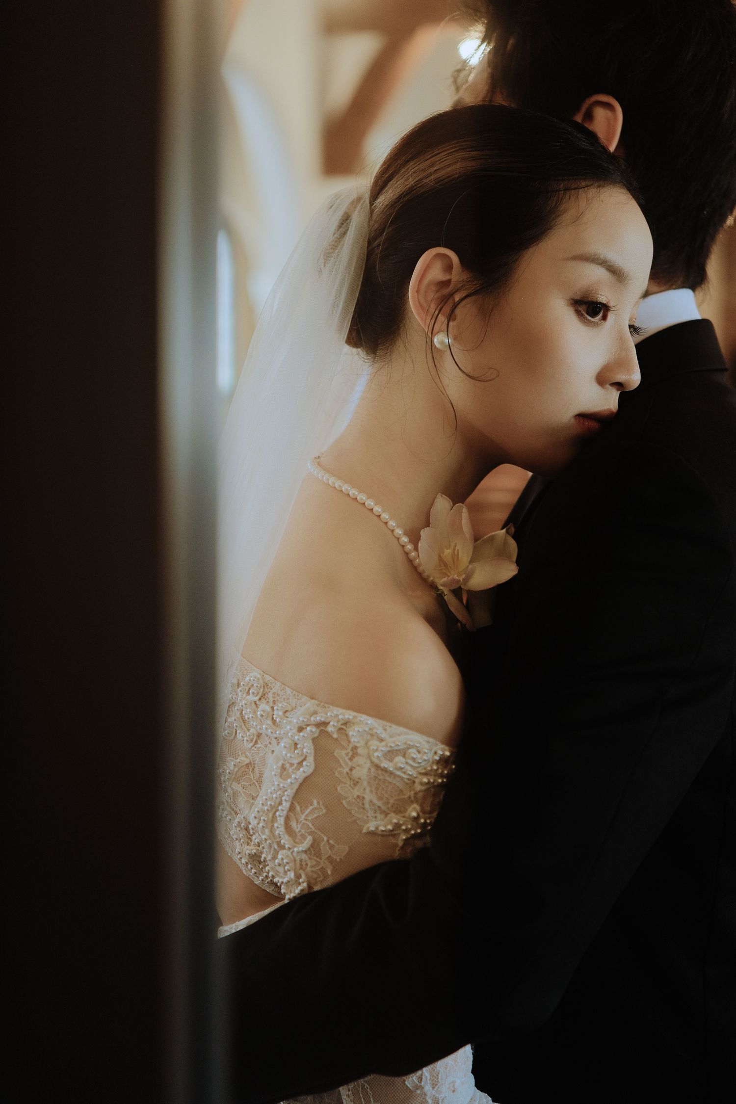 Bride in off-shoulder lace gown with pearl necklace embraces groom in a dark church.