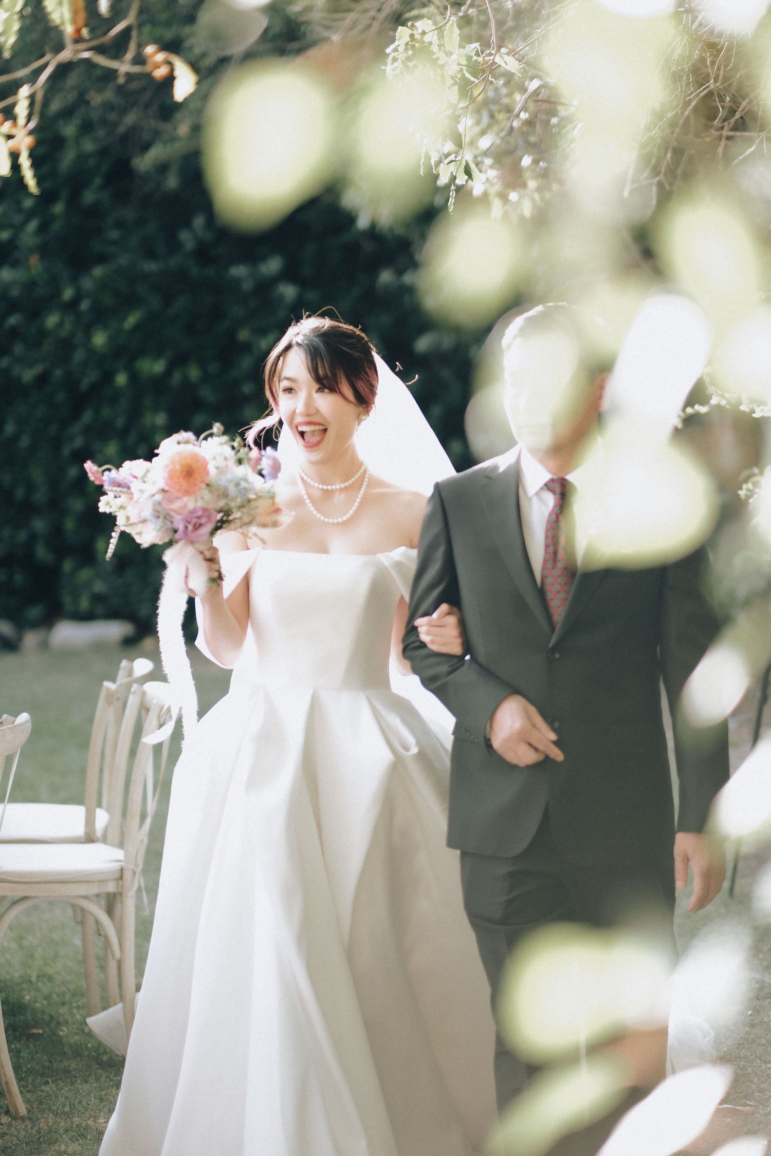 Smiling bride in off-shoulder white gown walks down aisle holding floral bouquet.