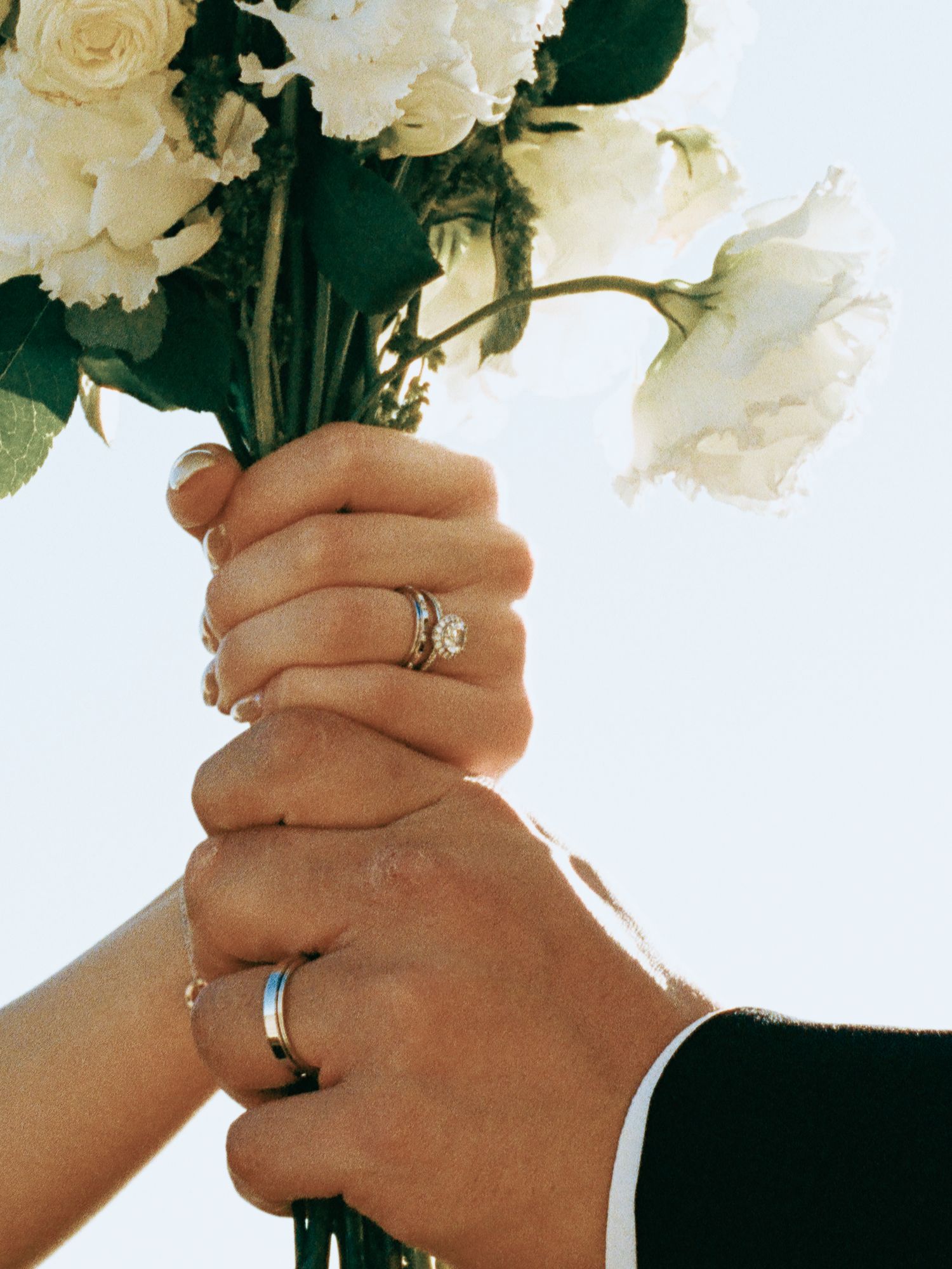 Couple holding white floral wedding bouquet together, displaying engagement and wedding rings.