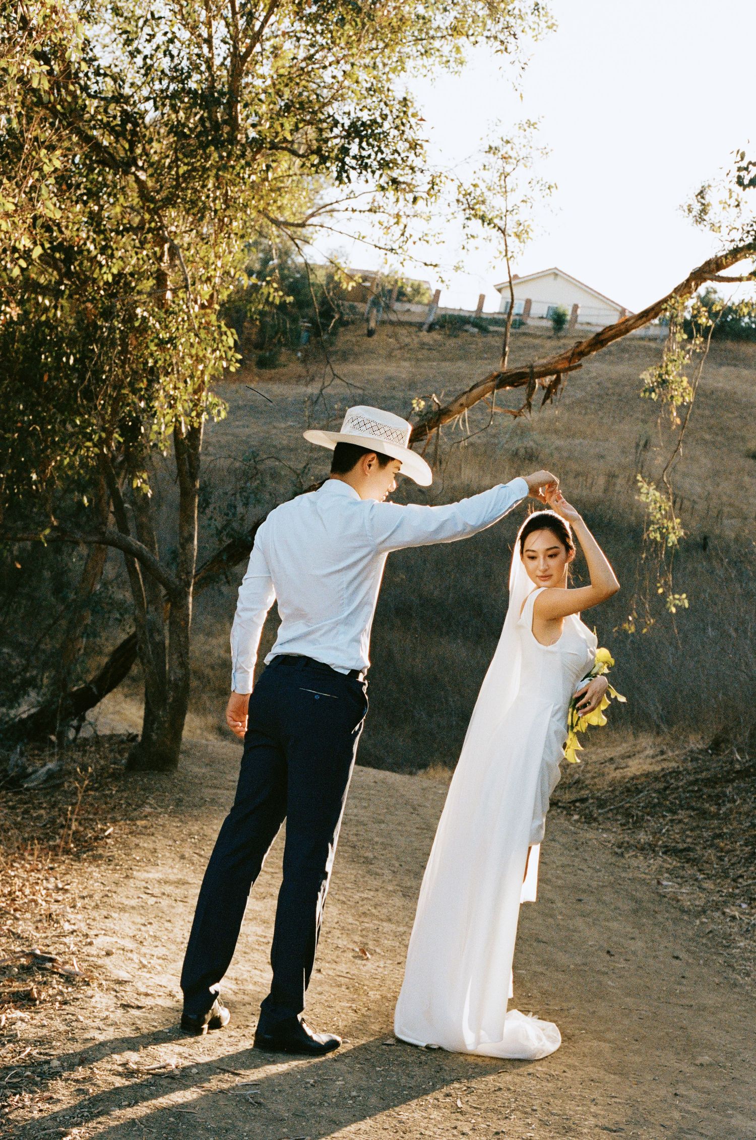 Bride and groom dancing on a sunlit dirt path, she in a white gown with veil, he in a cowboy hat.