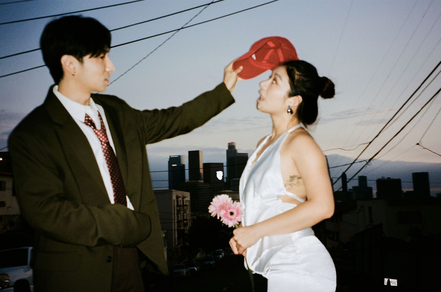 Couple sharing romantic moment on rooftop at dusk, man placing red cap on womans head, city skyline behind.