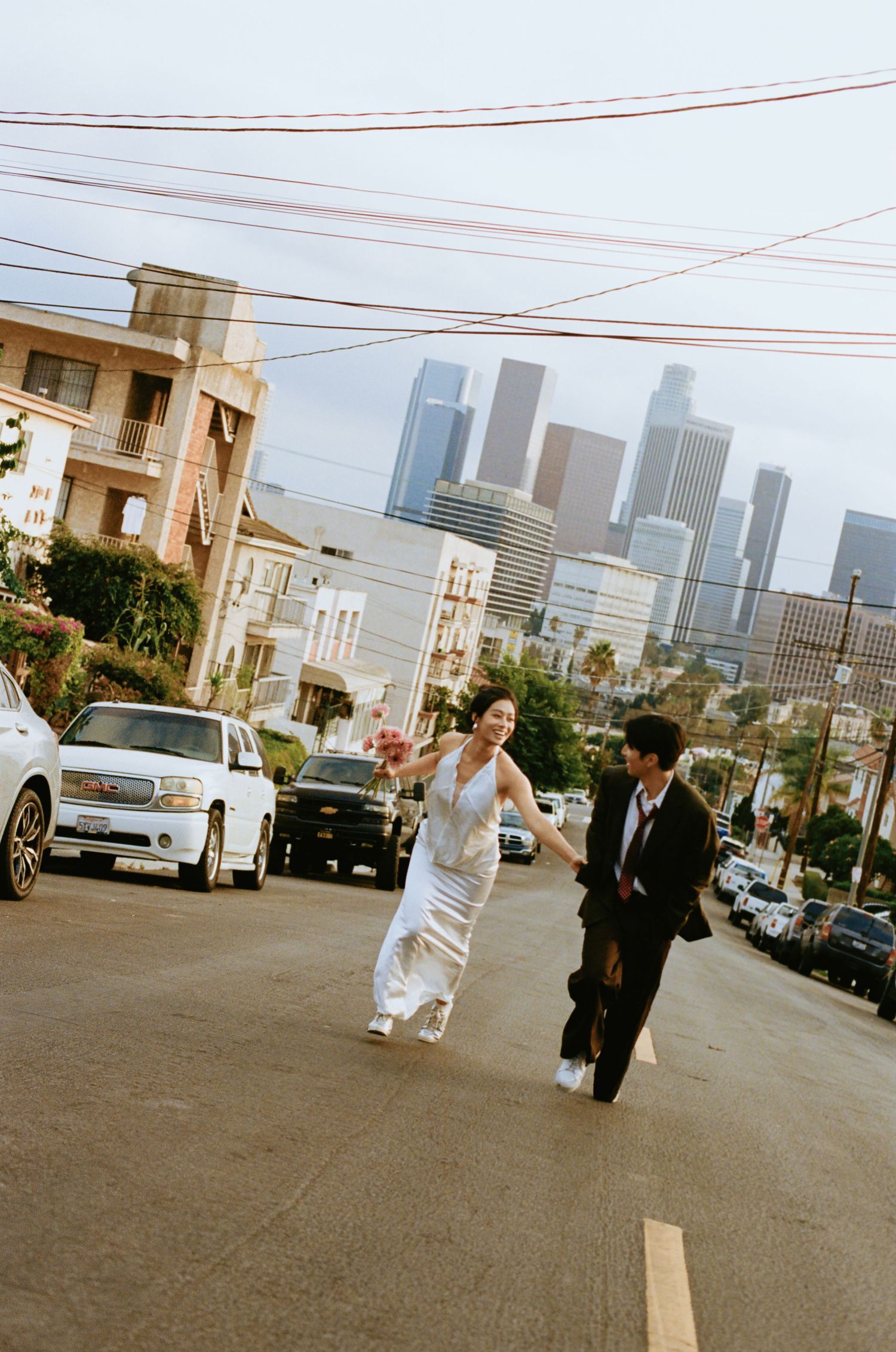 Bride in white dress and groom in suit run joyfully across a street with LA city skyline backdrop.