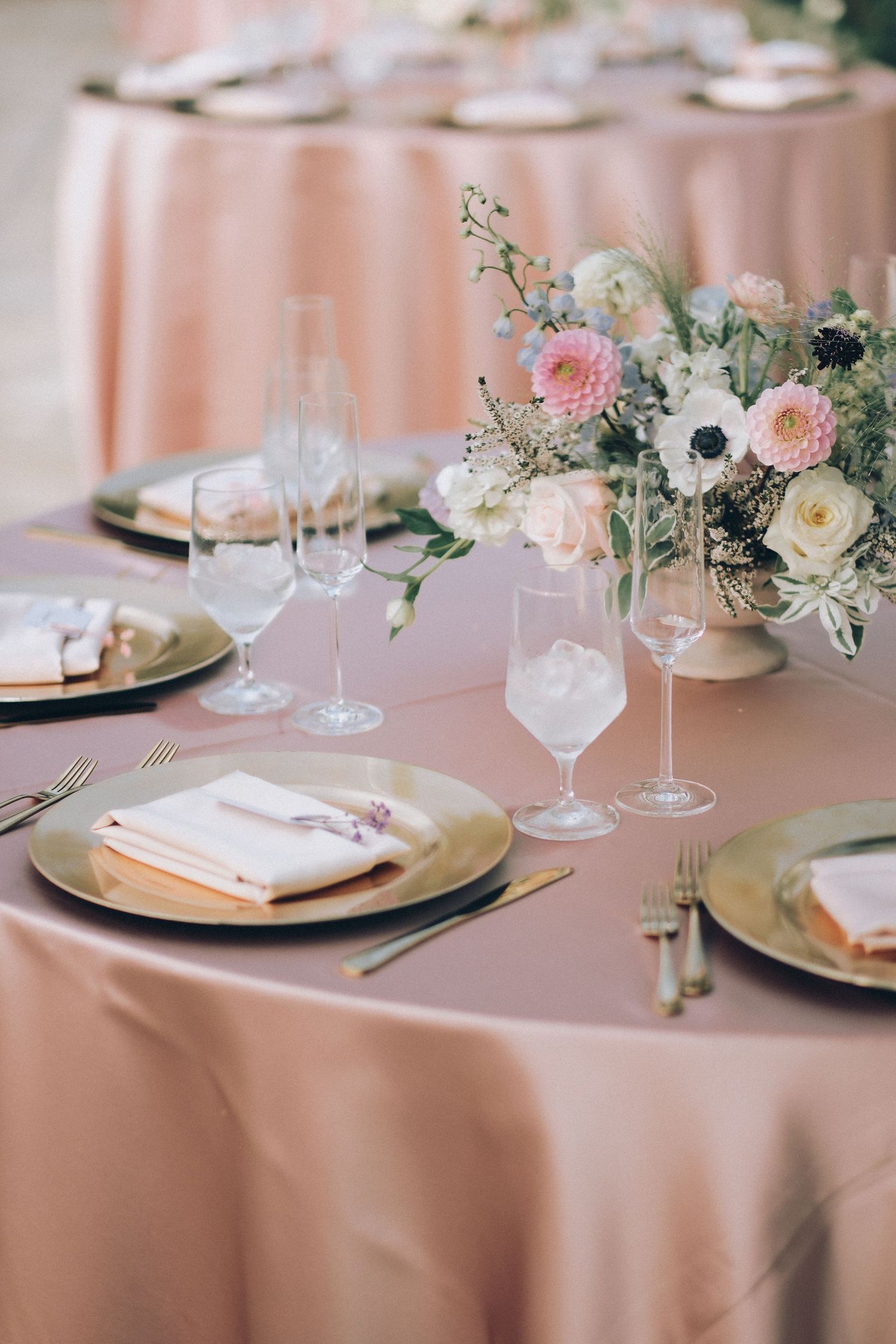 Wedding reception table set with gold charger plates, crystal glasses, pink napkins, and a pastel floral centerpiece.