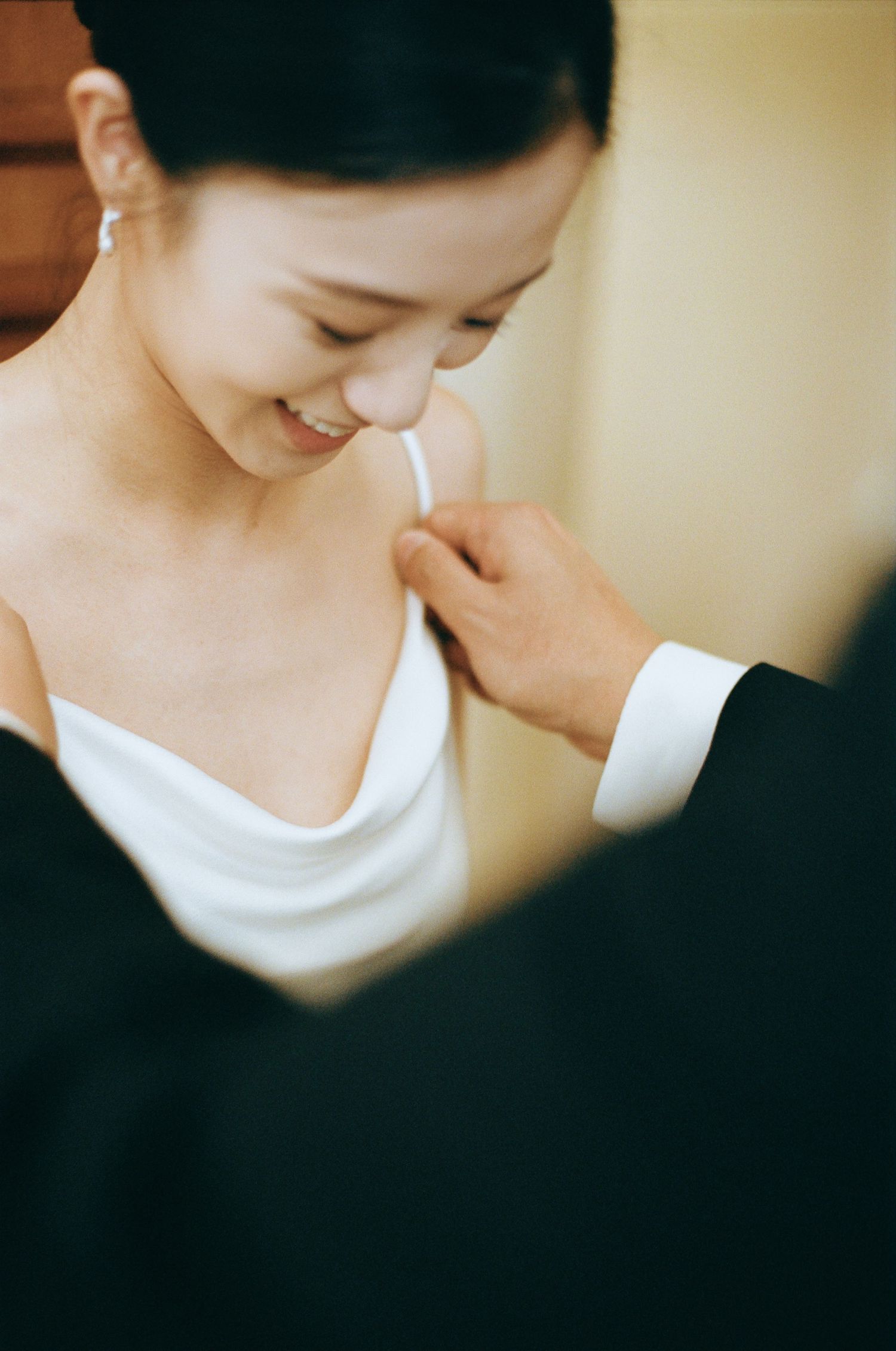 Smiling Asian bride in white gown getting dress strap adjusted by groom in black tuxedo.