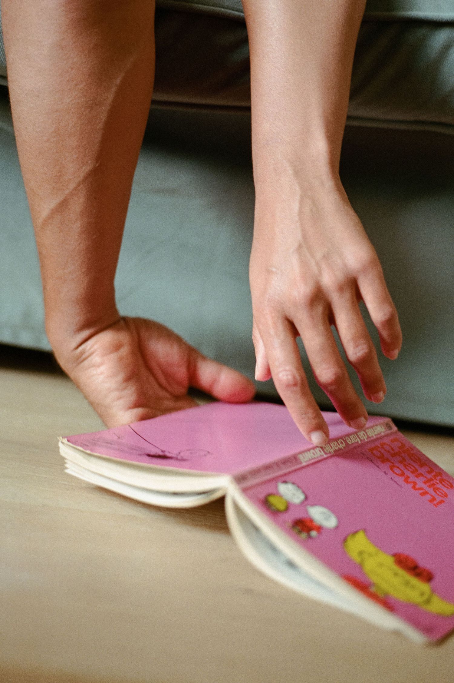 Close-up of hands browsing a pink book on a wooden floor near a grey sofa.
